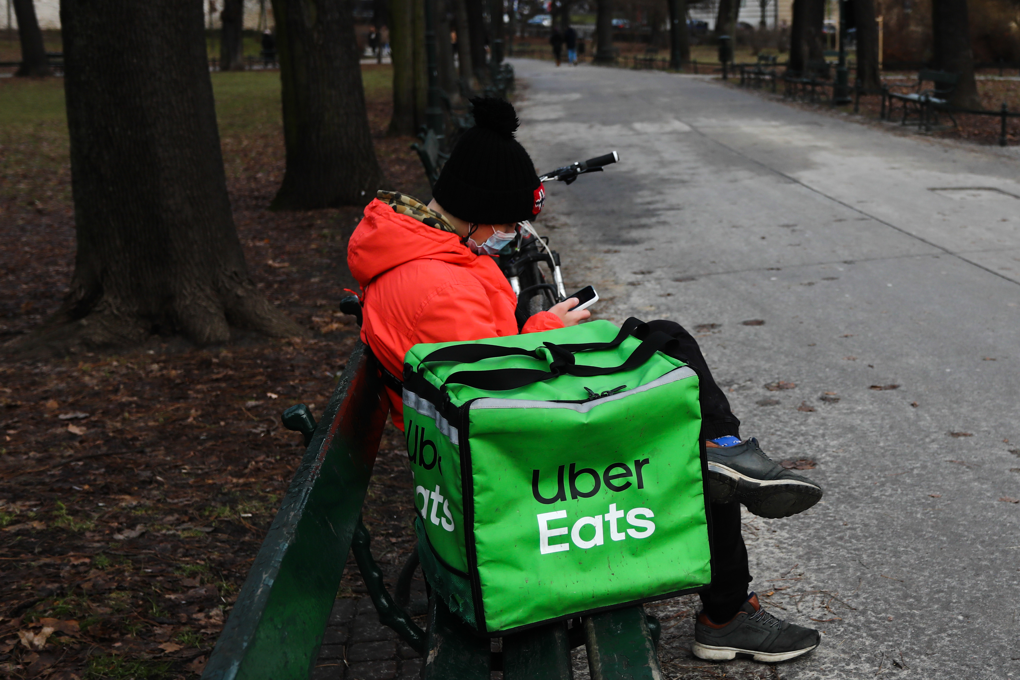 Person in orange jacket sits on a bench next to an insulated delivery bag with the UberEats logo.