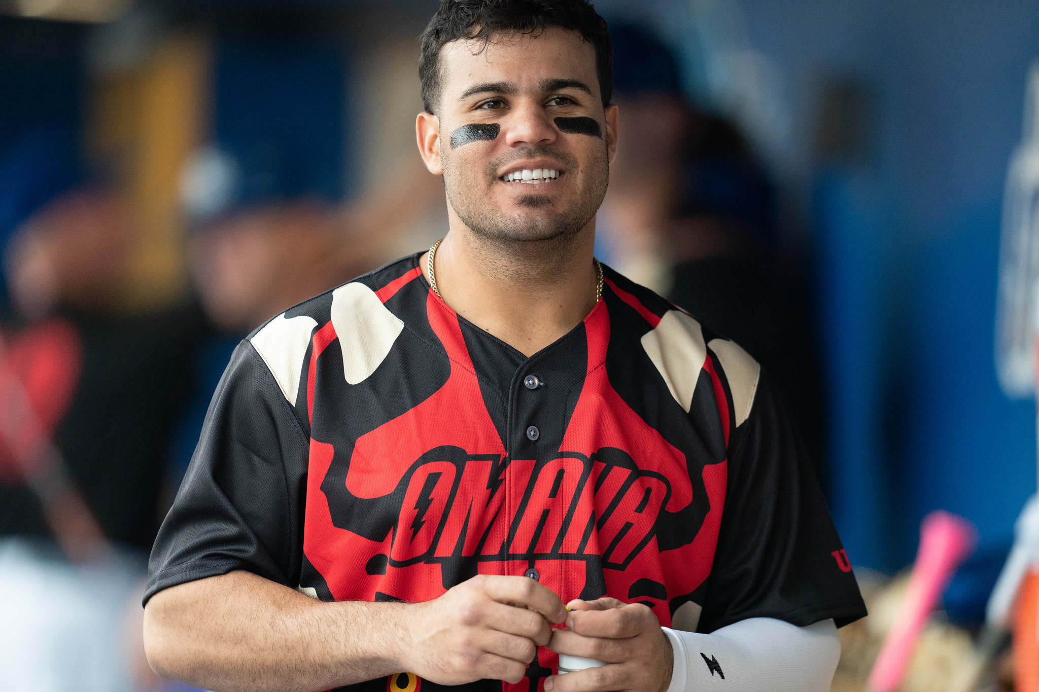 A baseball player with dark brown hair and eyeblack under both eyes standing in a dugout.