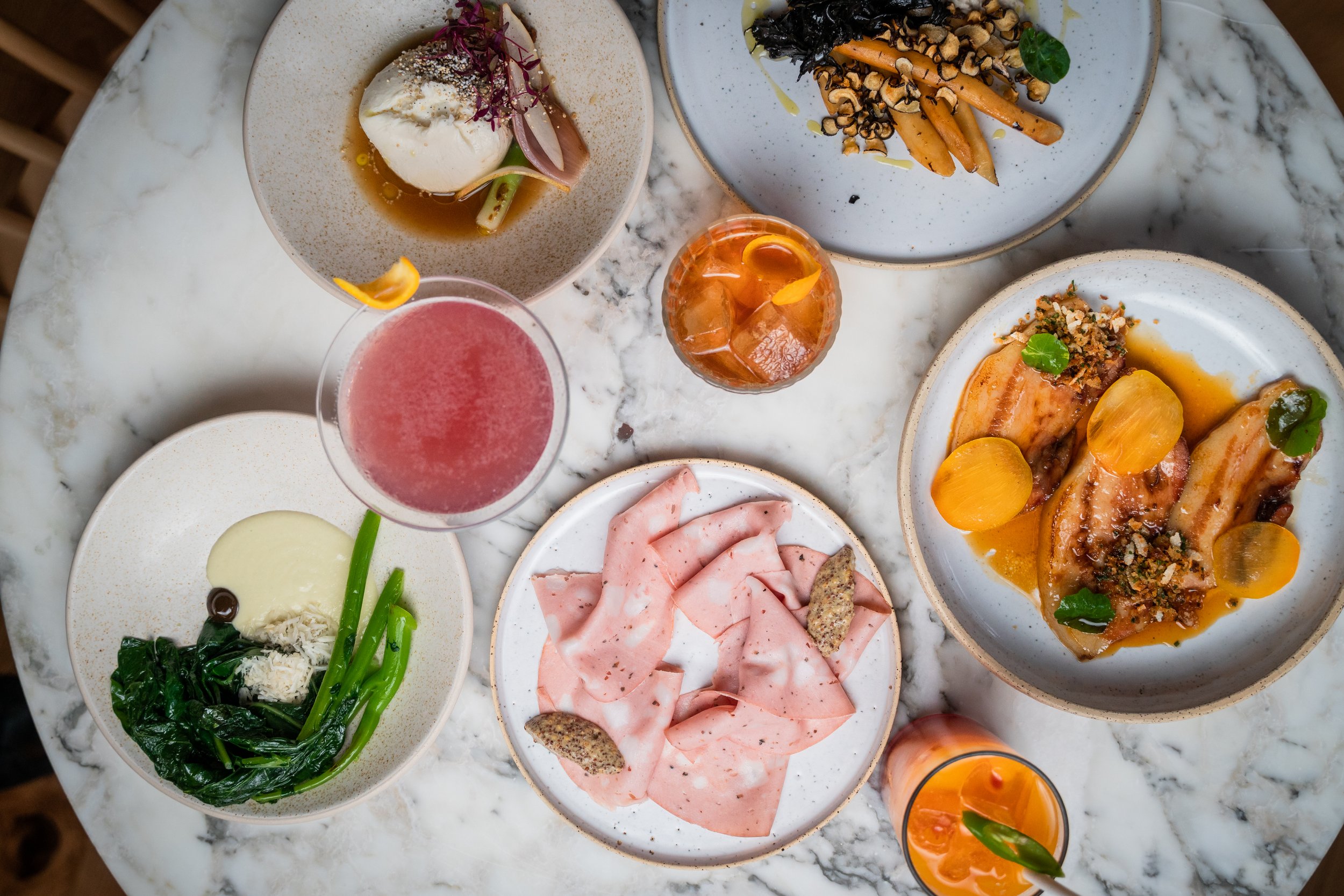 Plates of mortadella, charred broccoli, burrata, and roasted carrots on a marble tabletop