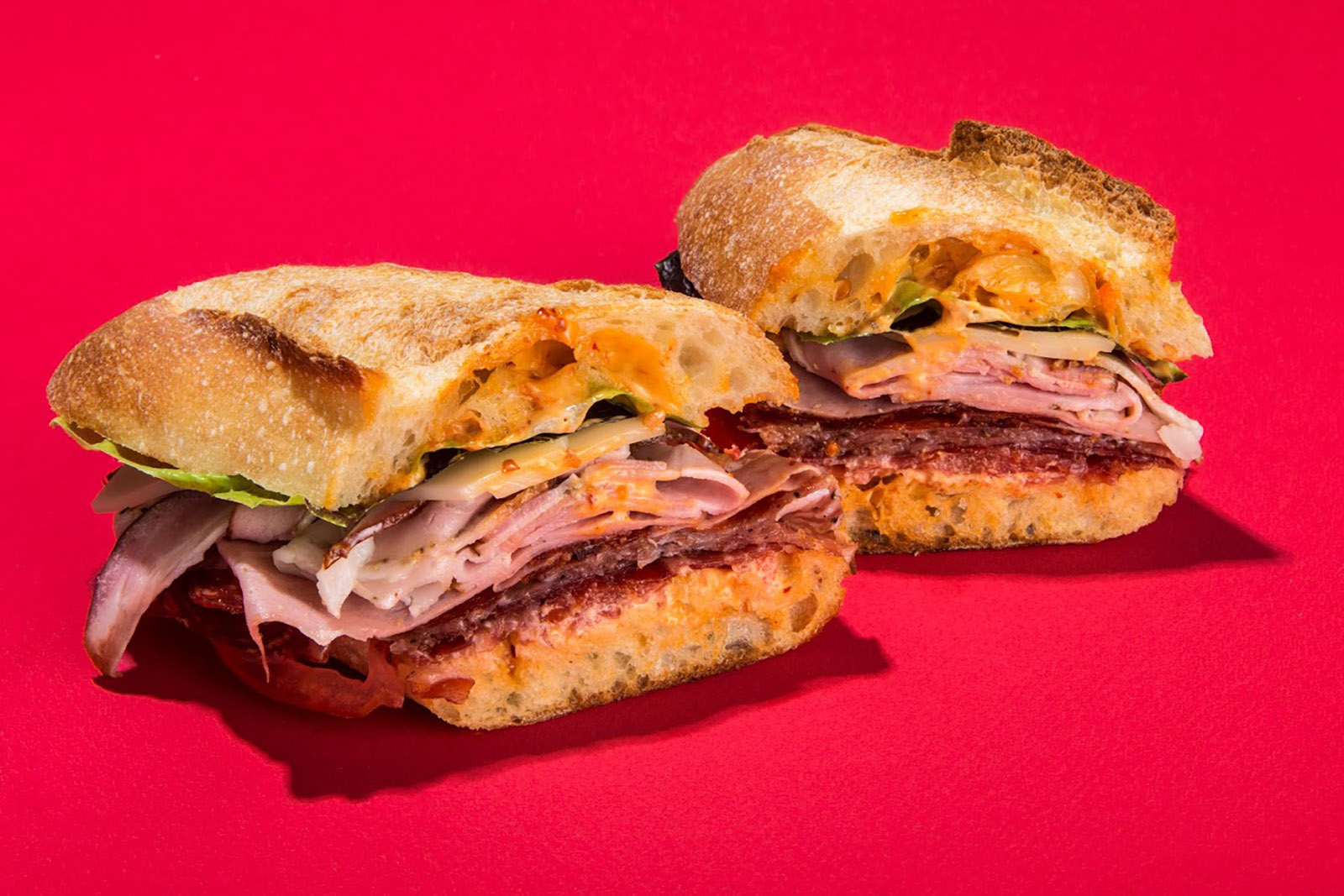 A sandwich filled with Italian meats, lettuce, and cheese in a baguette against a bright red background.