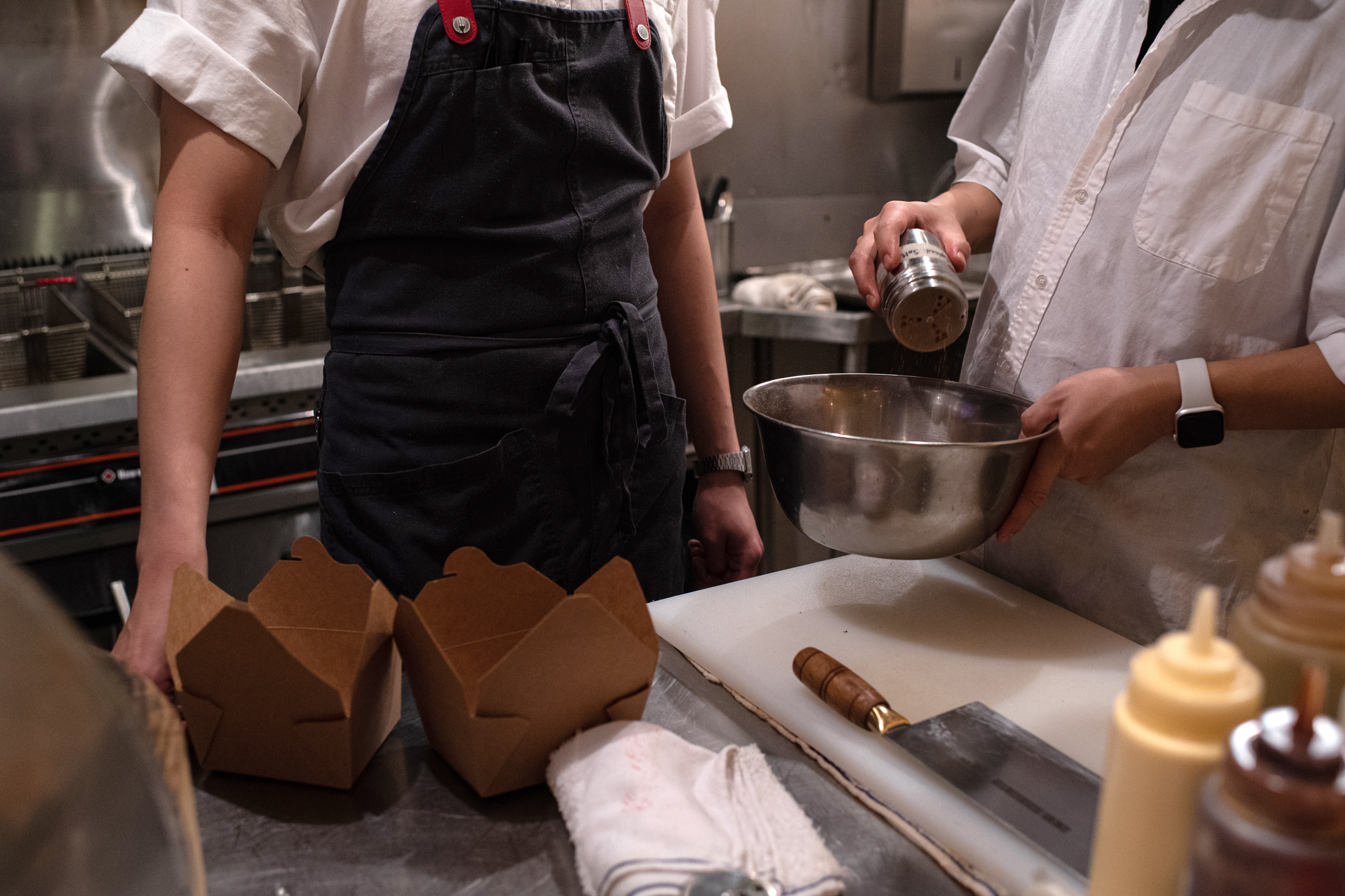 Two restaurant workers in a kitchen standing next to empty take-out containers; one figure is adding spices to a metal bowl.