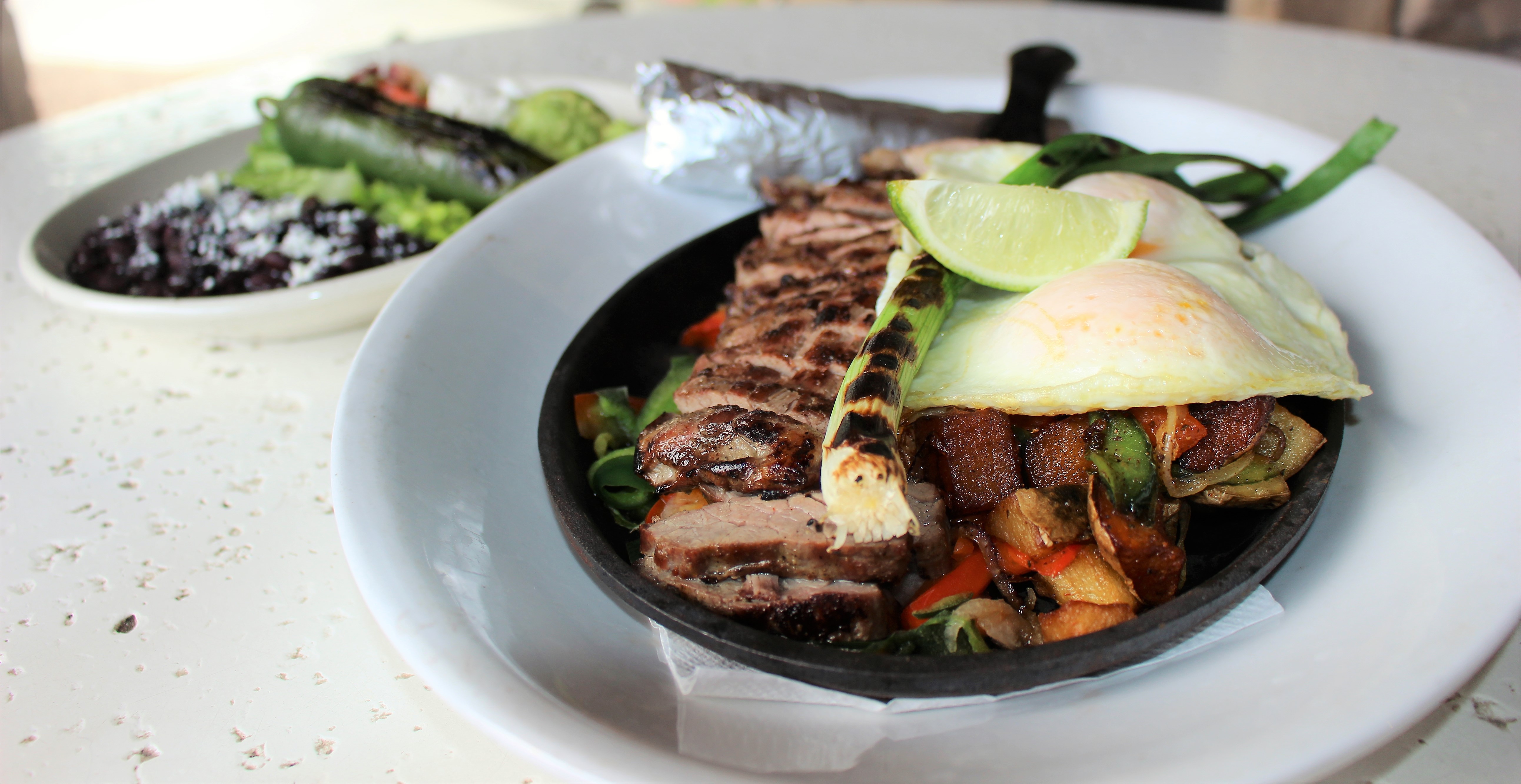 A skillet with steak, vegetables, a fried egg, and a lime on a white plate with another plate in the background with vegetables. 