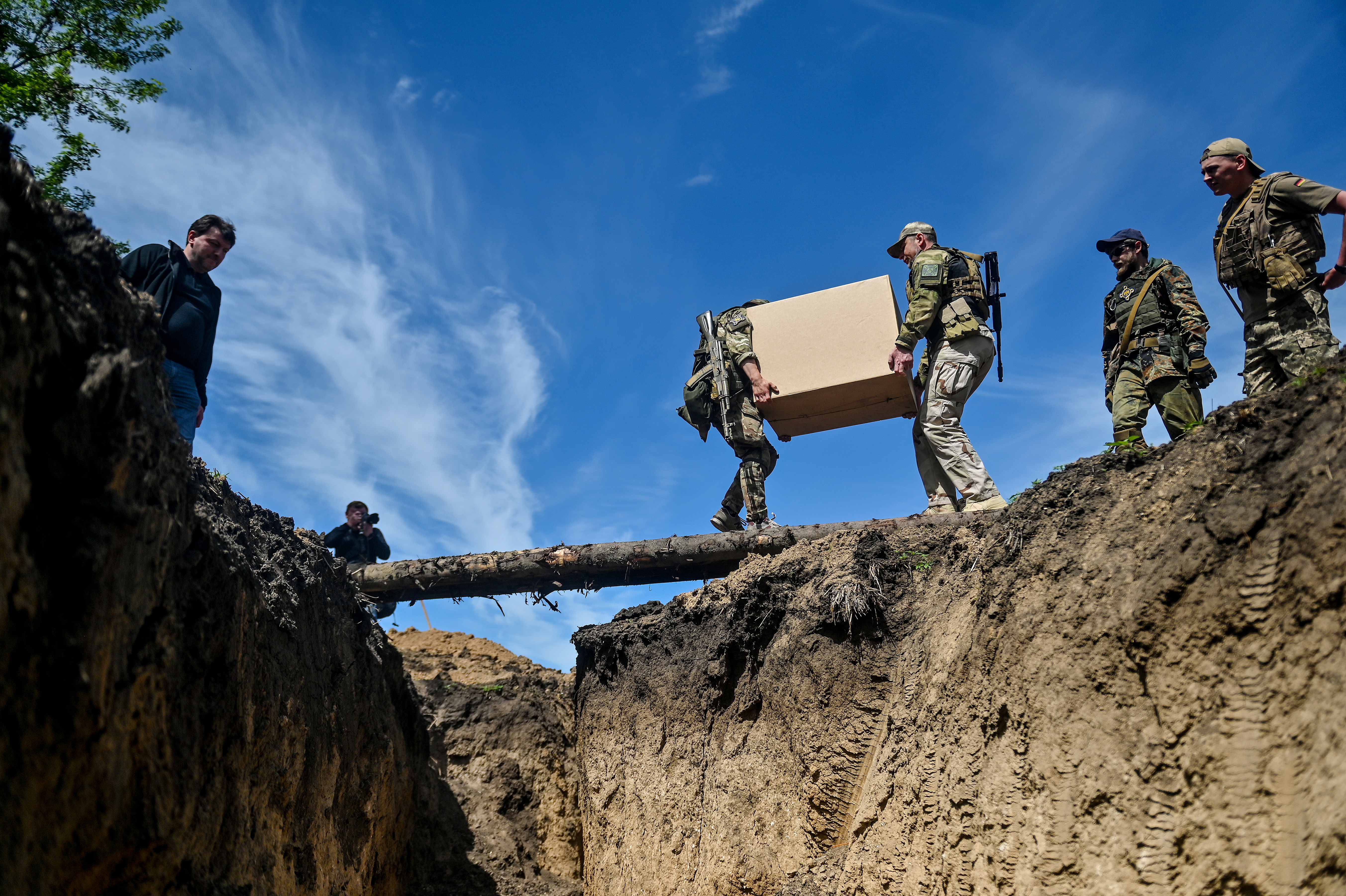 Under a bright blue sky, two soldiers in green and tan camouflage carry a tan box across a short bridge made out of logs that spans a trench. Other members of their squad look on.