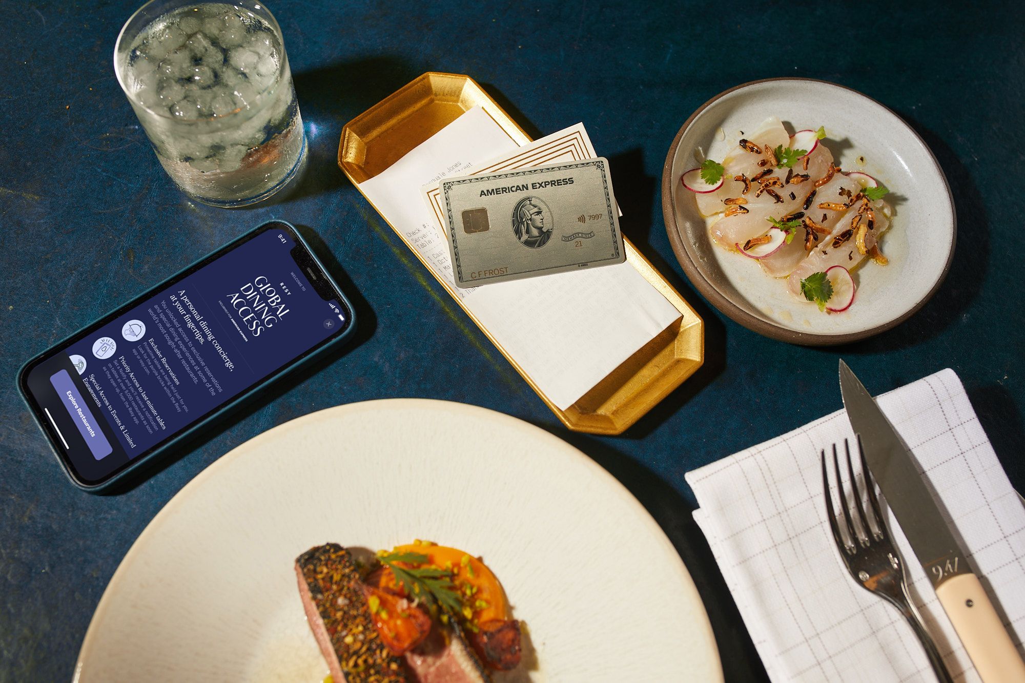 A Platinum American Express card sits adjacent a cocktail and an iPhone displaying Resy’s Global Dining Access Program; a plate of food sits below in the overhead shot