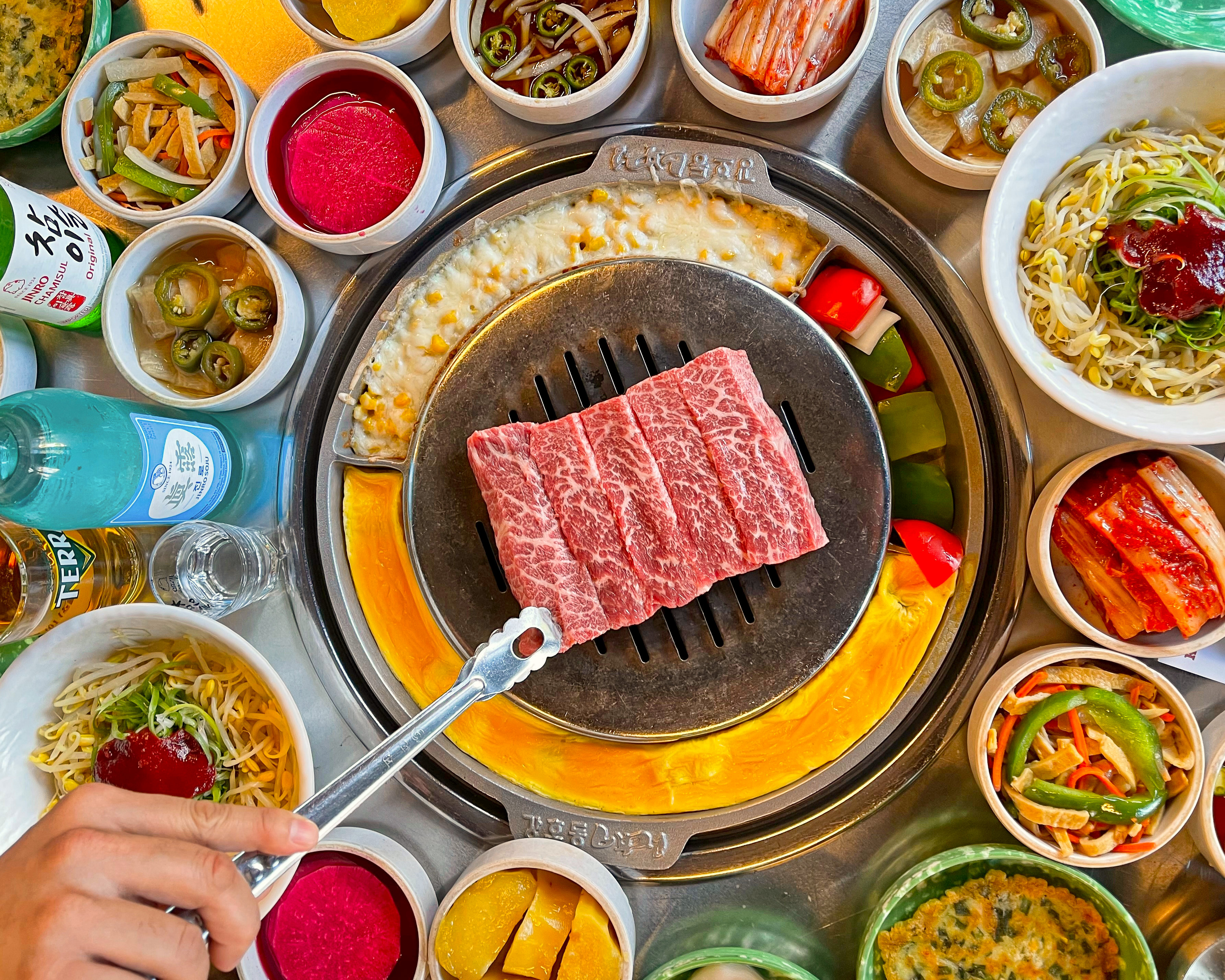 A cut of marbled beef on a grill, delicately handled with tongs. Around the grill are side dishes with sauces, pickled vegetables, and noodles.