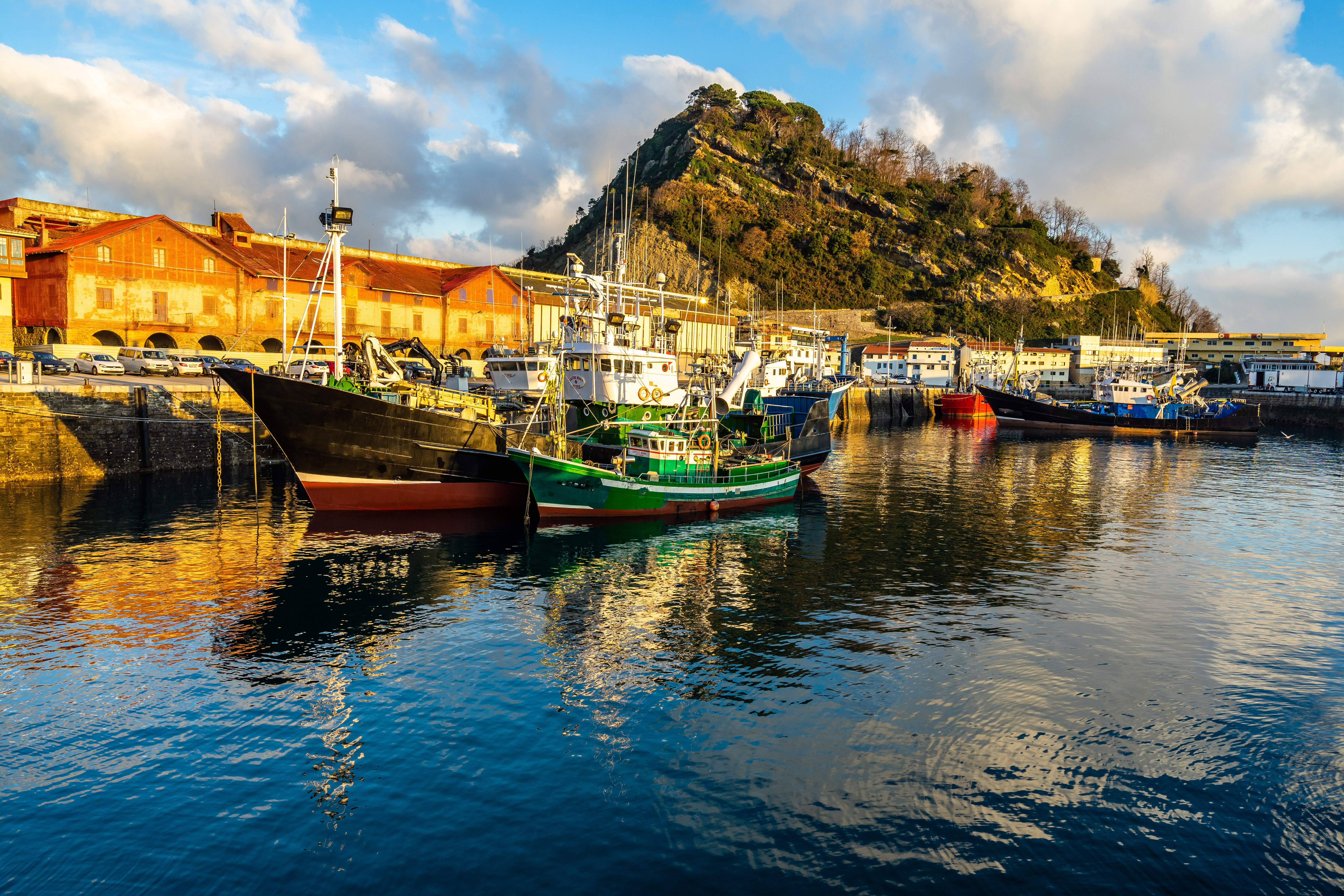 Boats moored in a harbor, with sunset-lit buildings and a large mountain in the background
