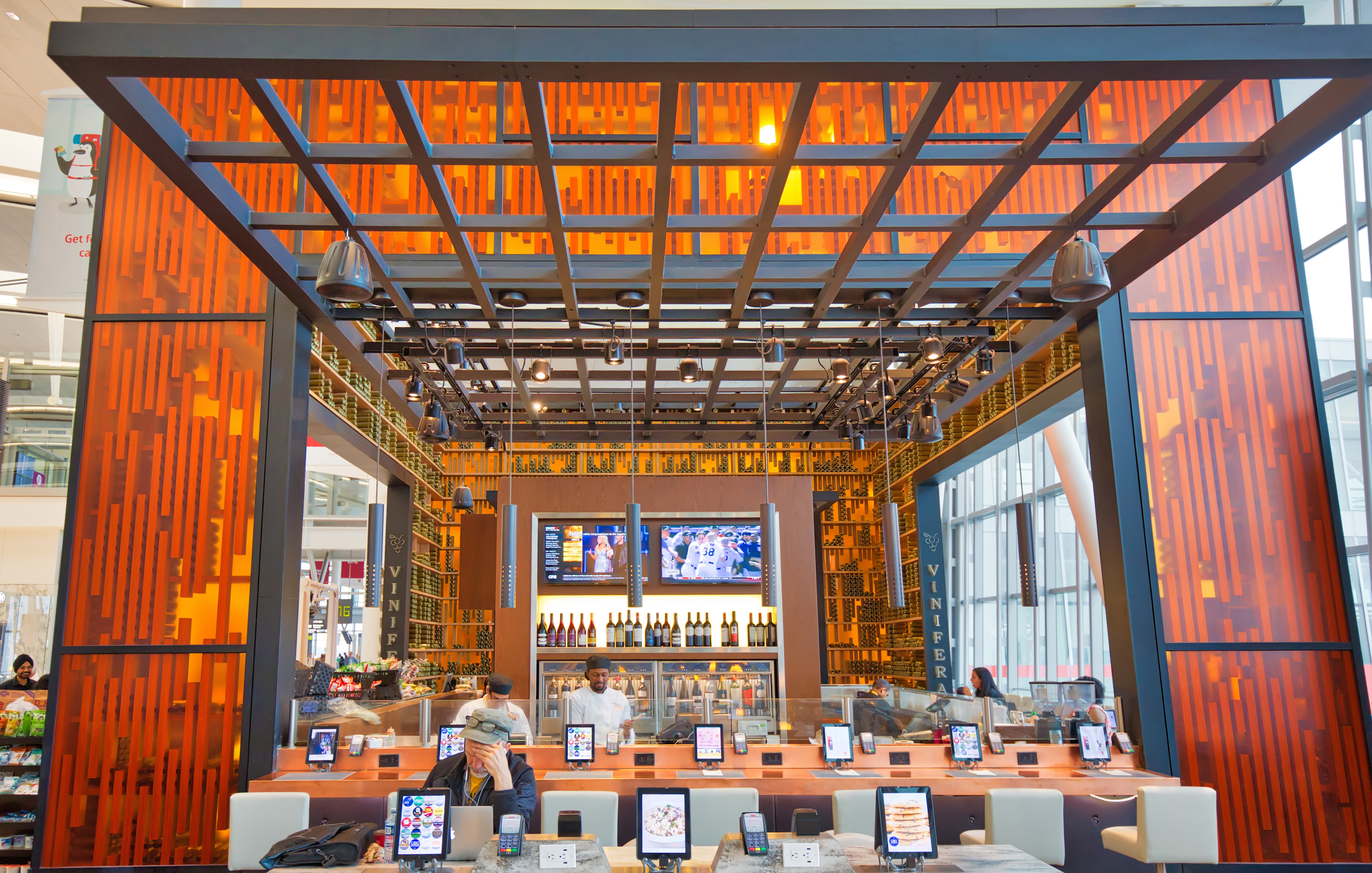 A large, open bar inside an airport hall. The restaurant is decorated with large orange panels. Customers sit at tables outfitted with tablets for ordering