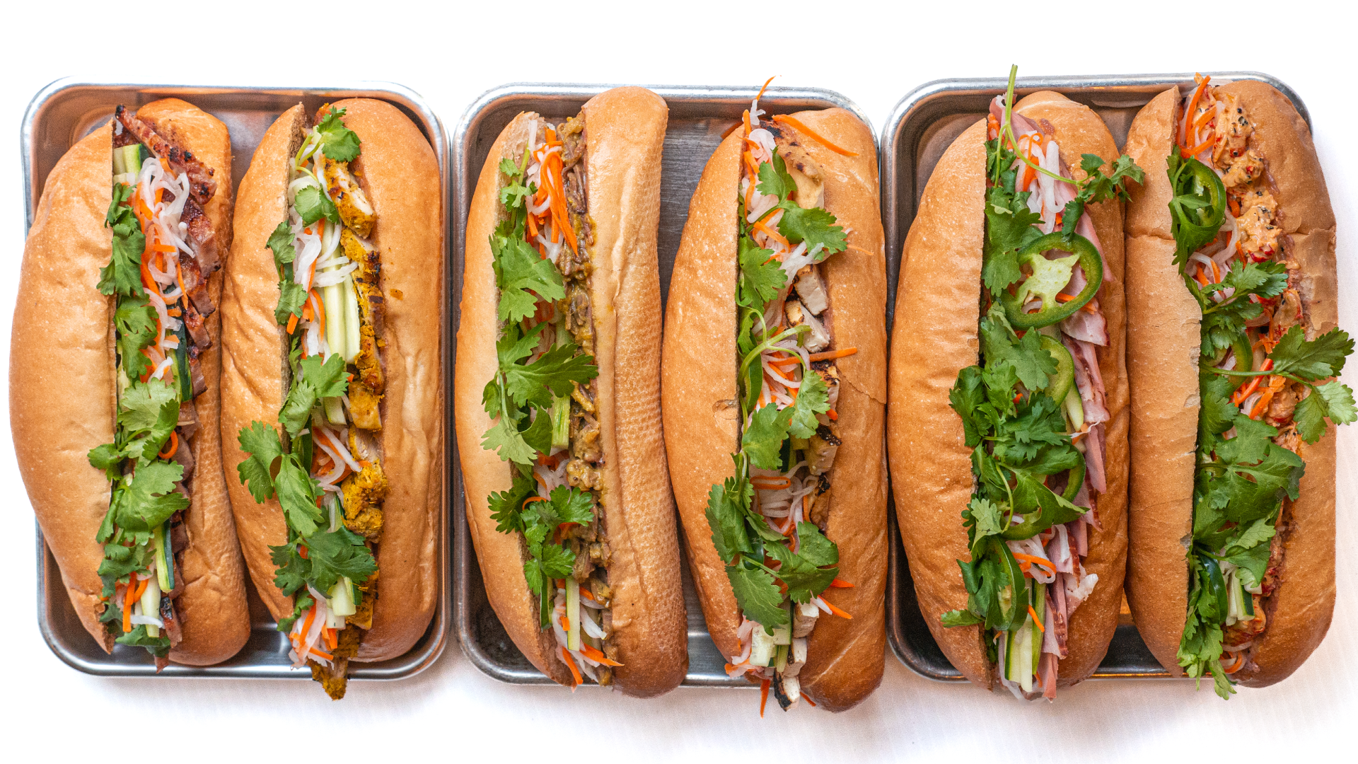 Overhead view of six Cambodian sandwiches on long sub rolls, arranged on small metal trays on a white background.