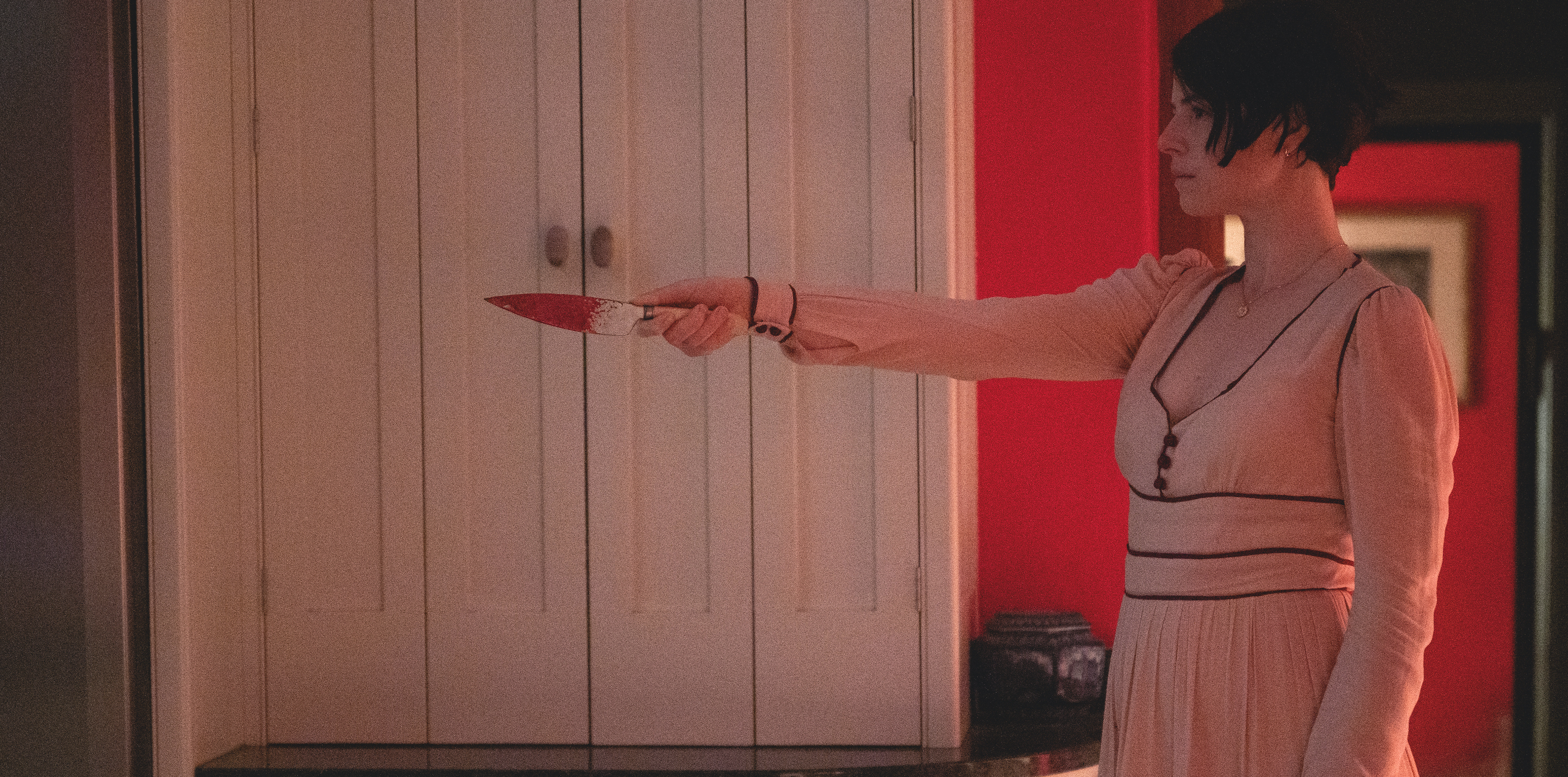 Jessie Buckley in a long pink dress, holding up a bloody knife in Men