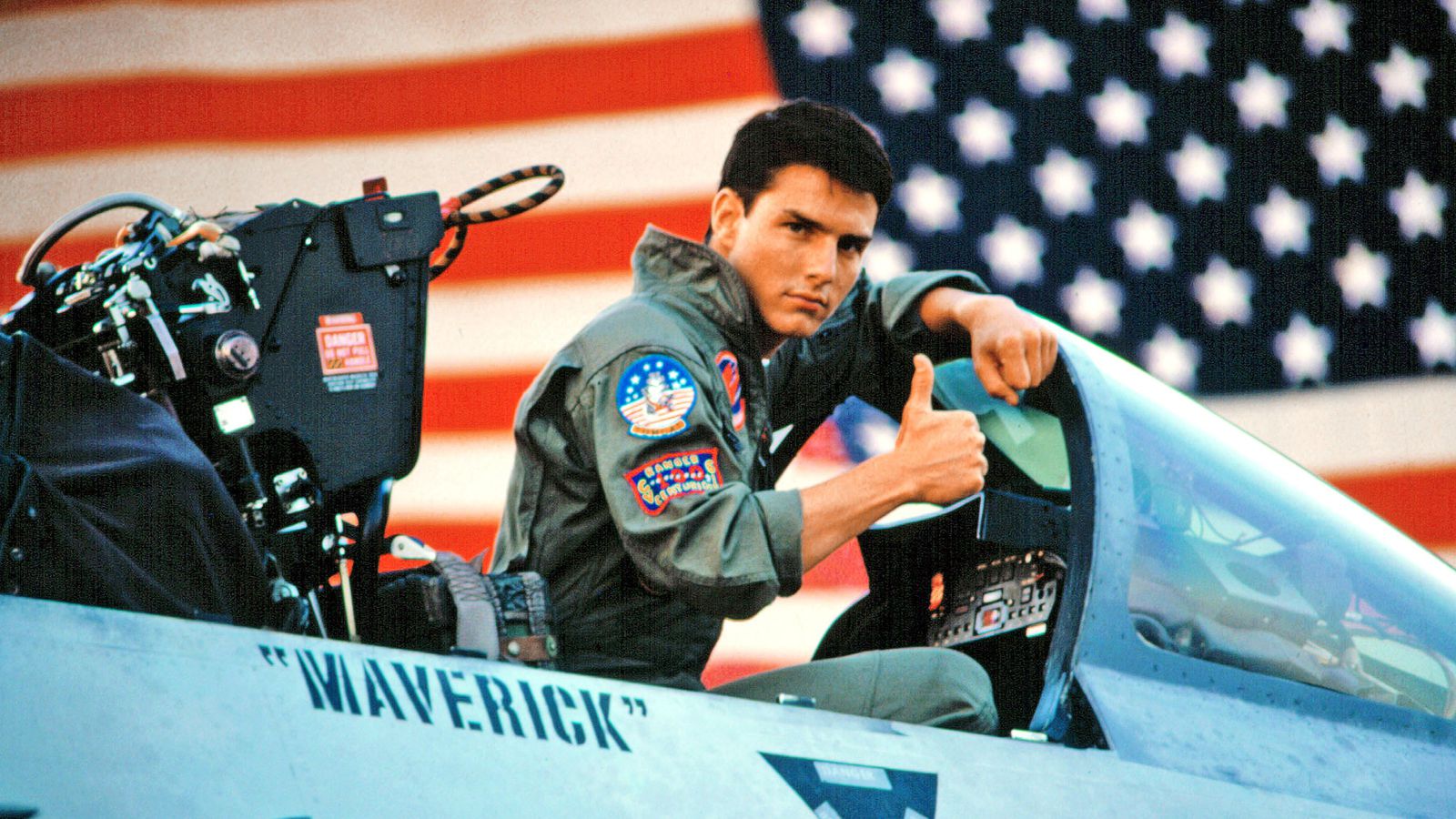 Tom Cruise gives a thumbs-up from the cockpit of a fighter jet, set against the backdrop of an American flag.