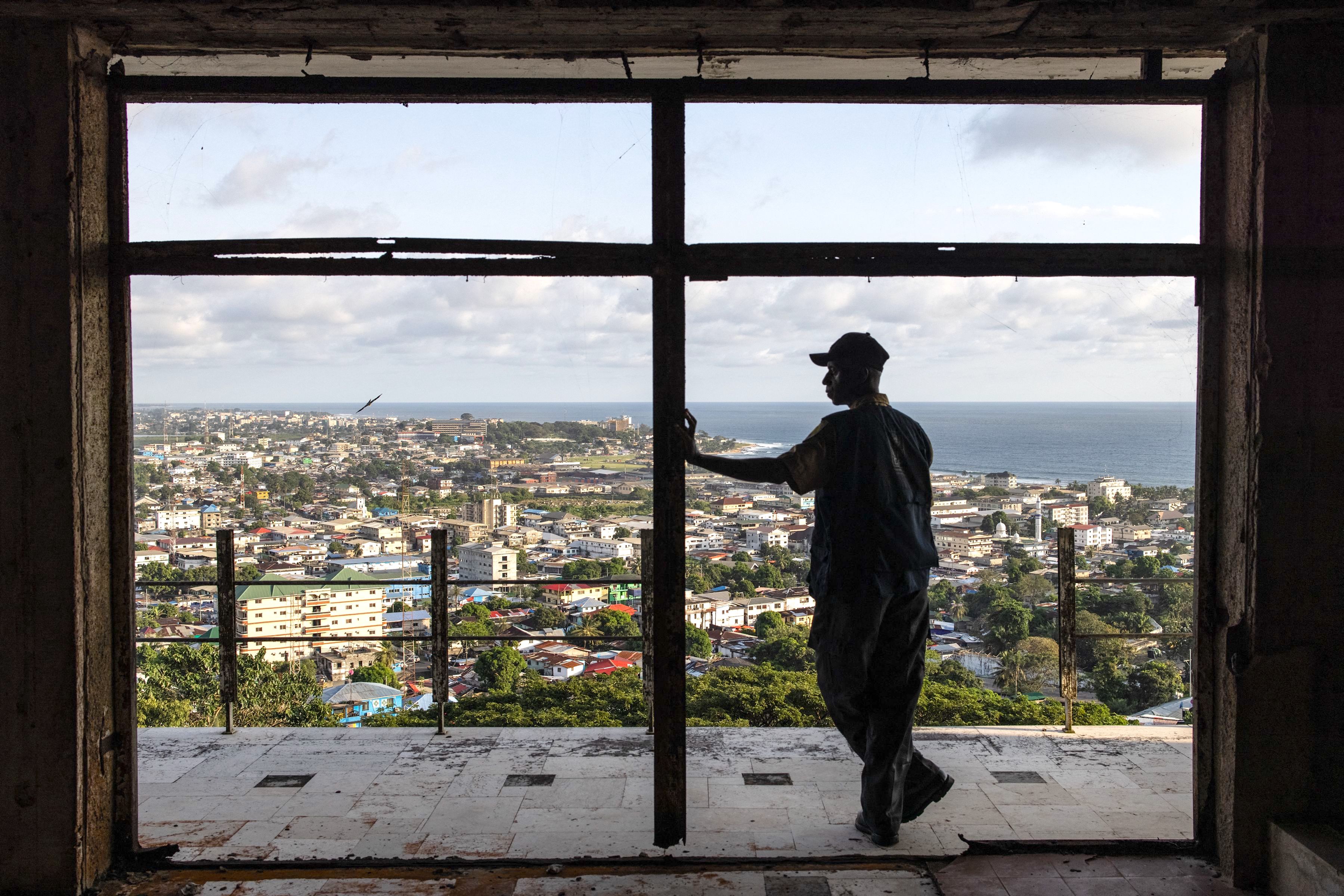 A person standing in the balcony opening of a derelict hotel overlooking a coastal city.