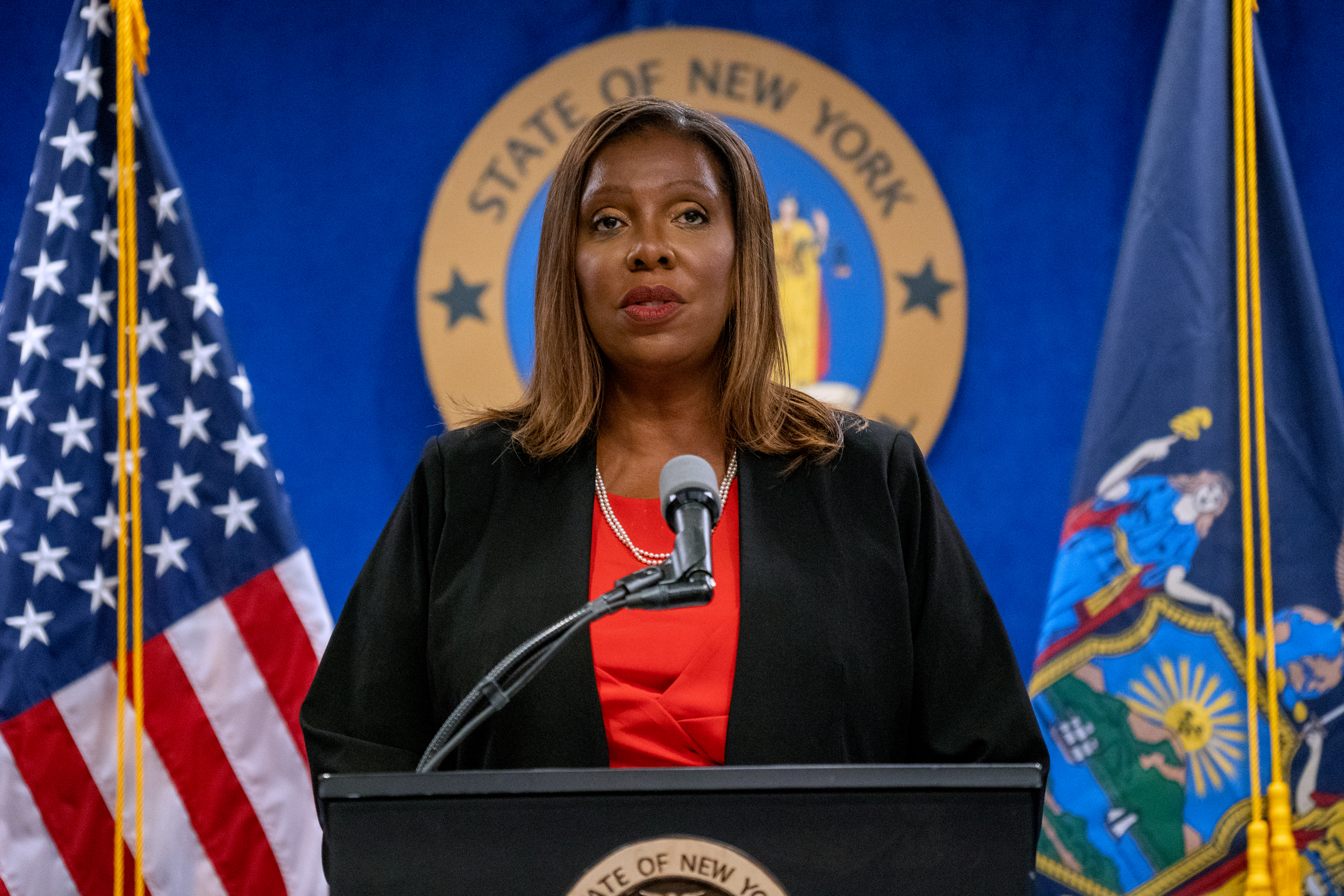 New York Attorney General Letitia James standing at a lectern and speaking into a microphone, with the seal of the state of New York behind her and flags on either side.