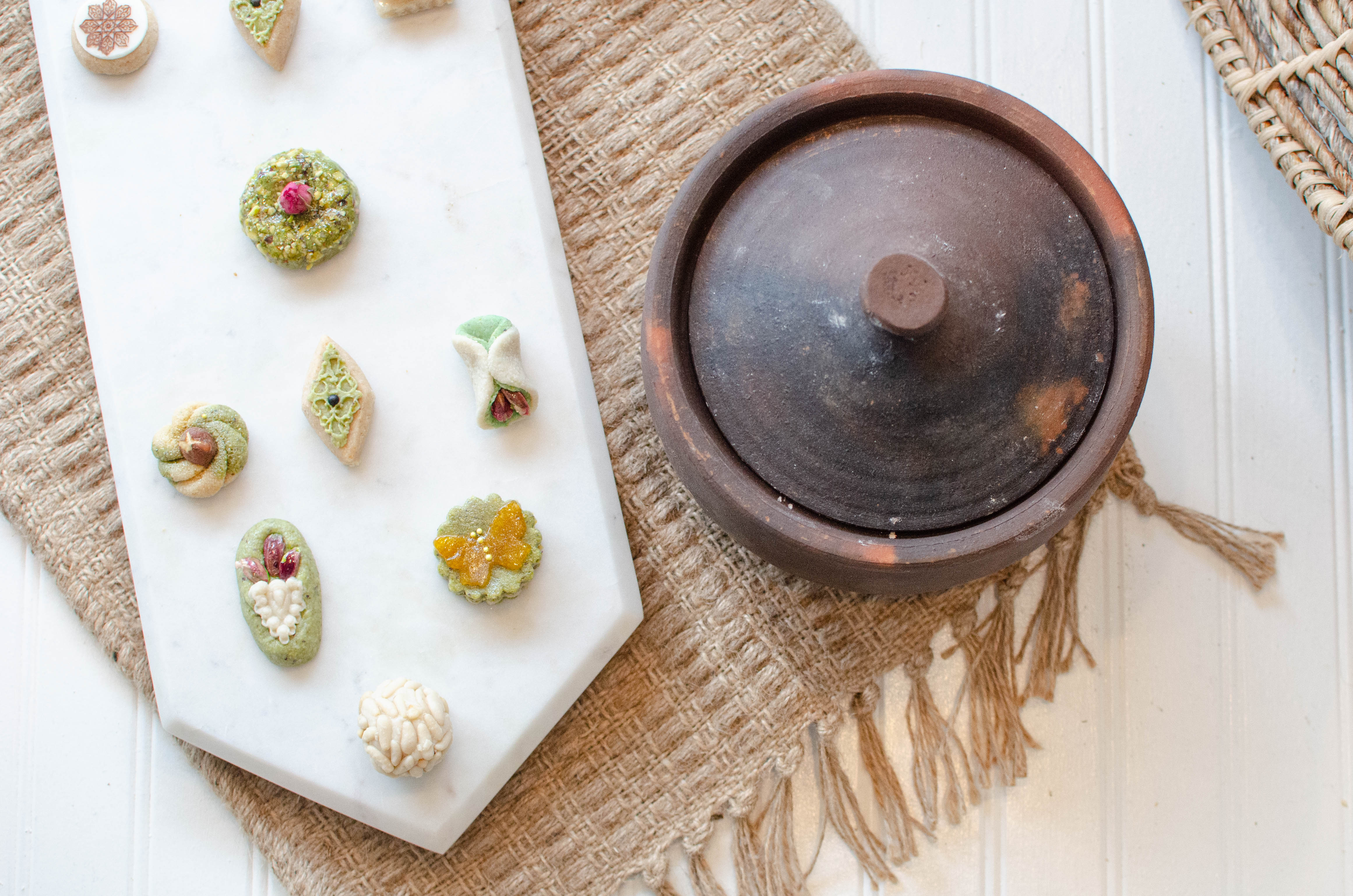 Small artistic-looking pastries made of pistachios, pinenuts, dried roses, and more are displayed on a white marble board.