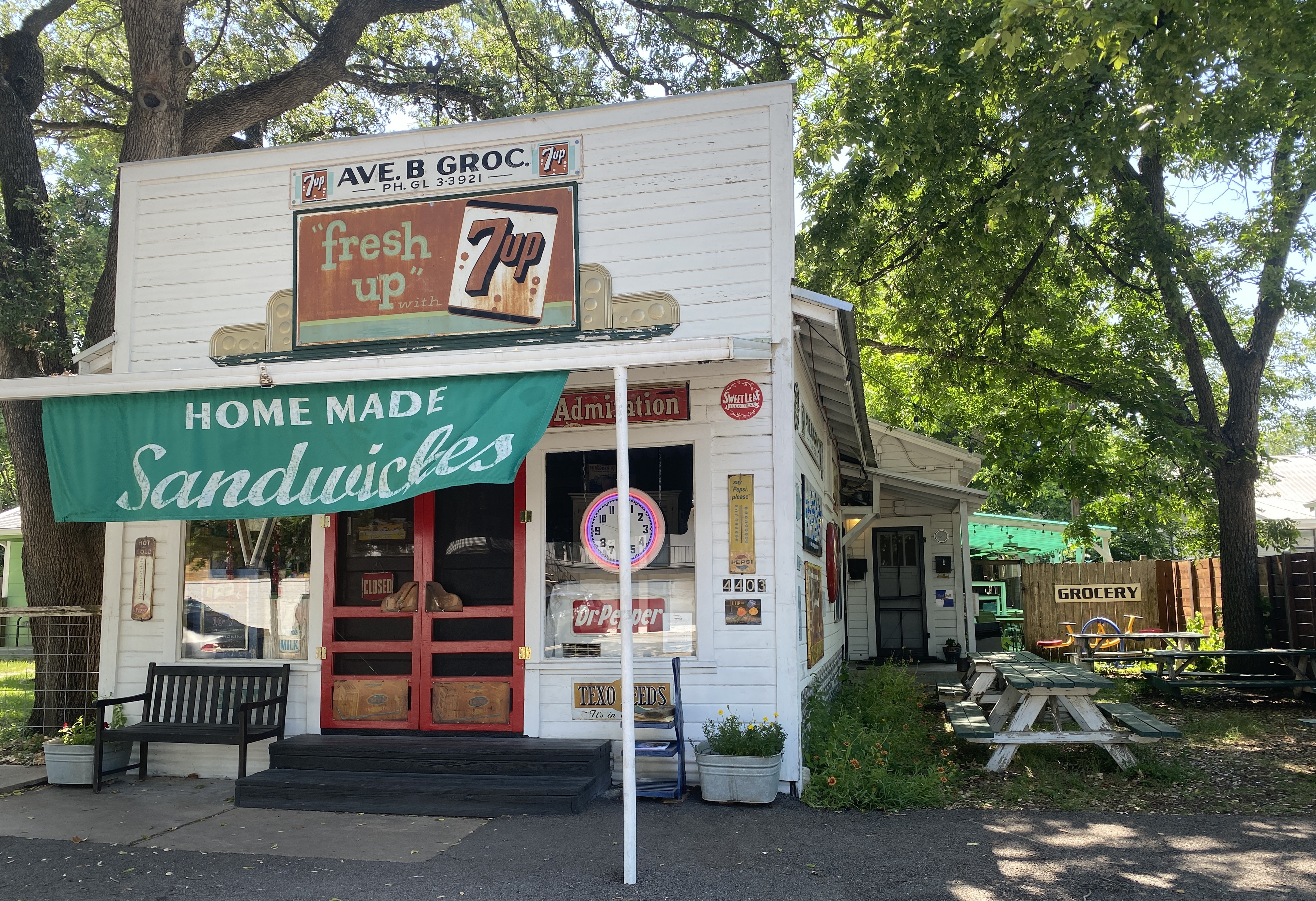 A white old-timey store front with antique soda signs, a banner that says Homemade Sandwiches, and picnic tables to the side and trees in the background