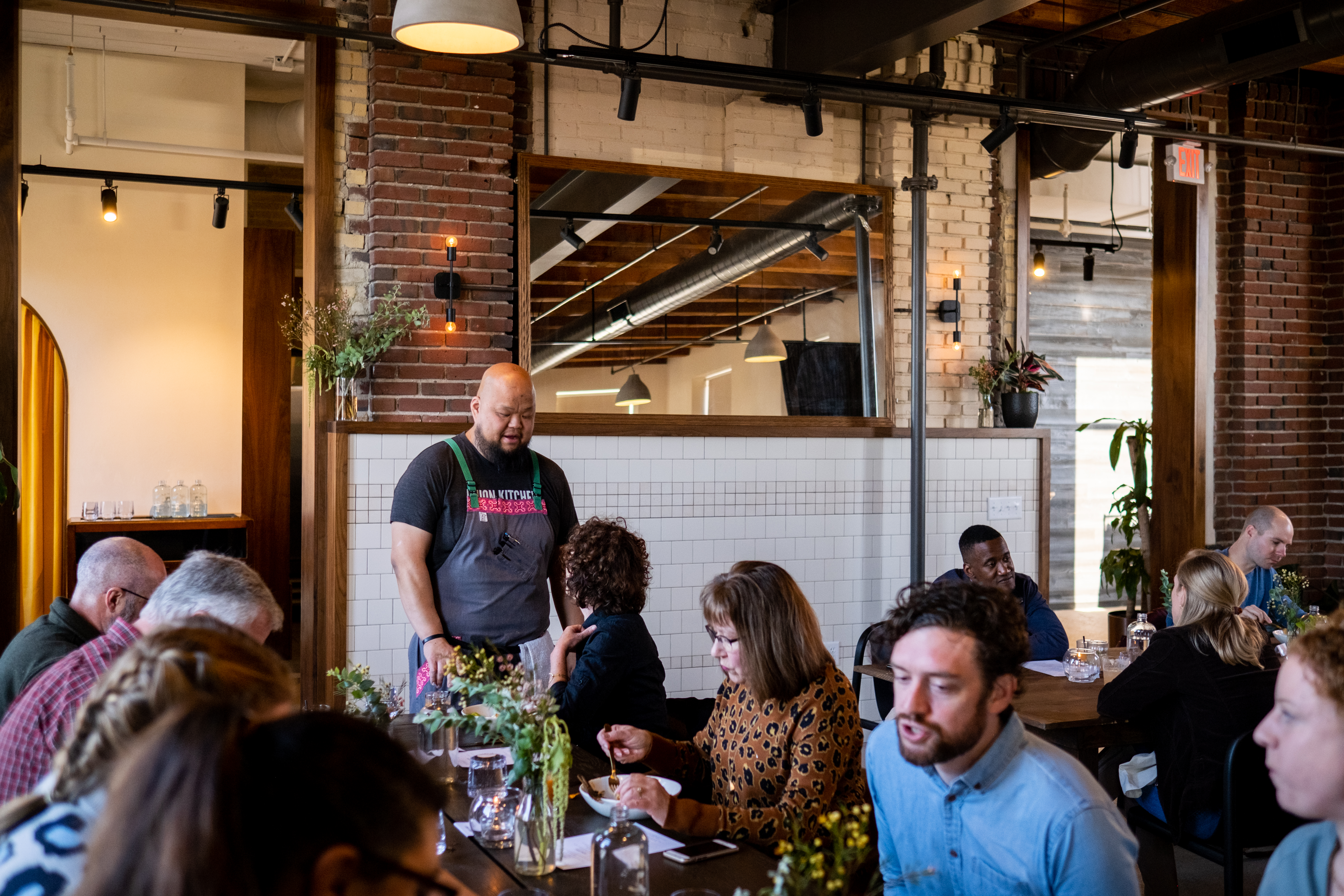 Chef Yia Vang stands at a table in the background wearing a black T-Shirt and grey apron. Diners are seated and dining in the foreground; behind Vang is a brick wall with large windows and track lighting. 