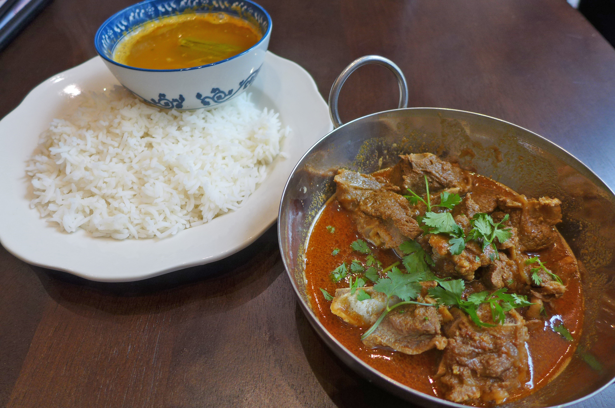 A metal wok of dark red meat curry on the bottom right, with a plate of rice and cup of soup on the upper left.