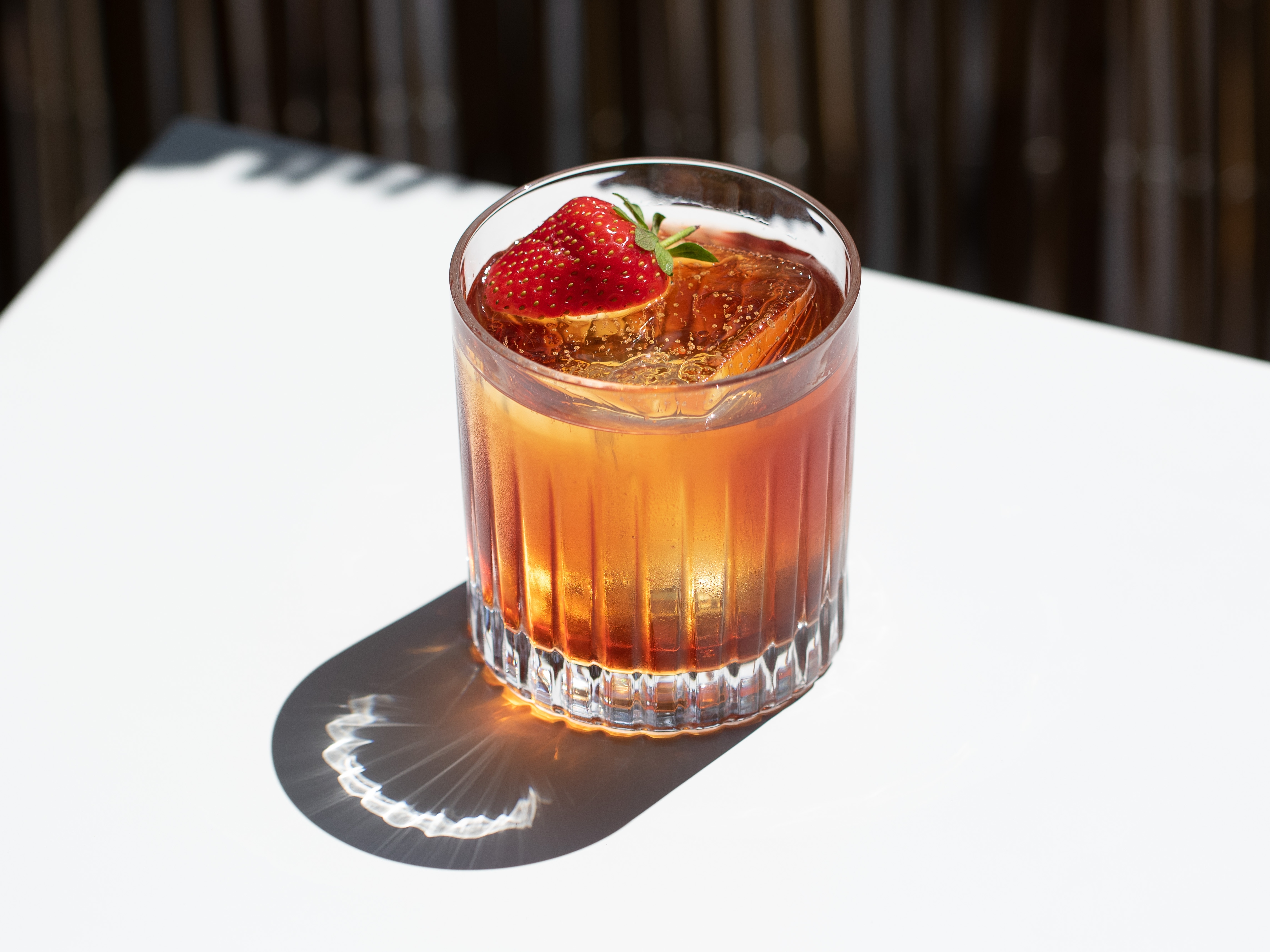 A mezcal Negroni basks in the sunlight on a white table with large ice cubs and a floating strawberry.