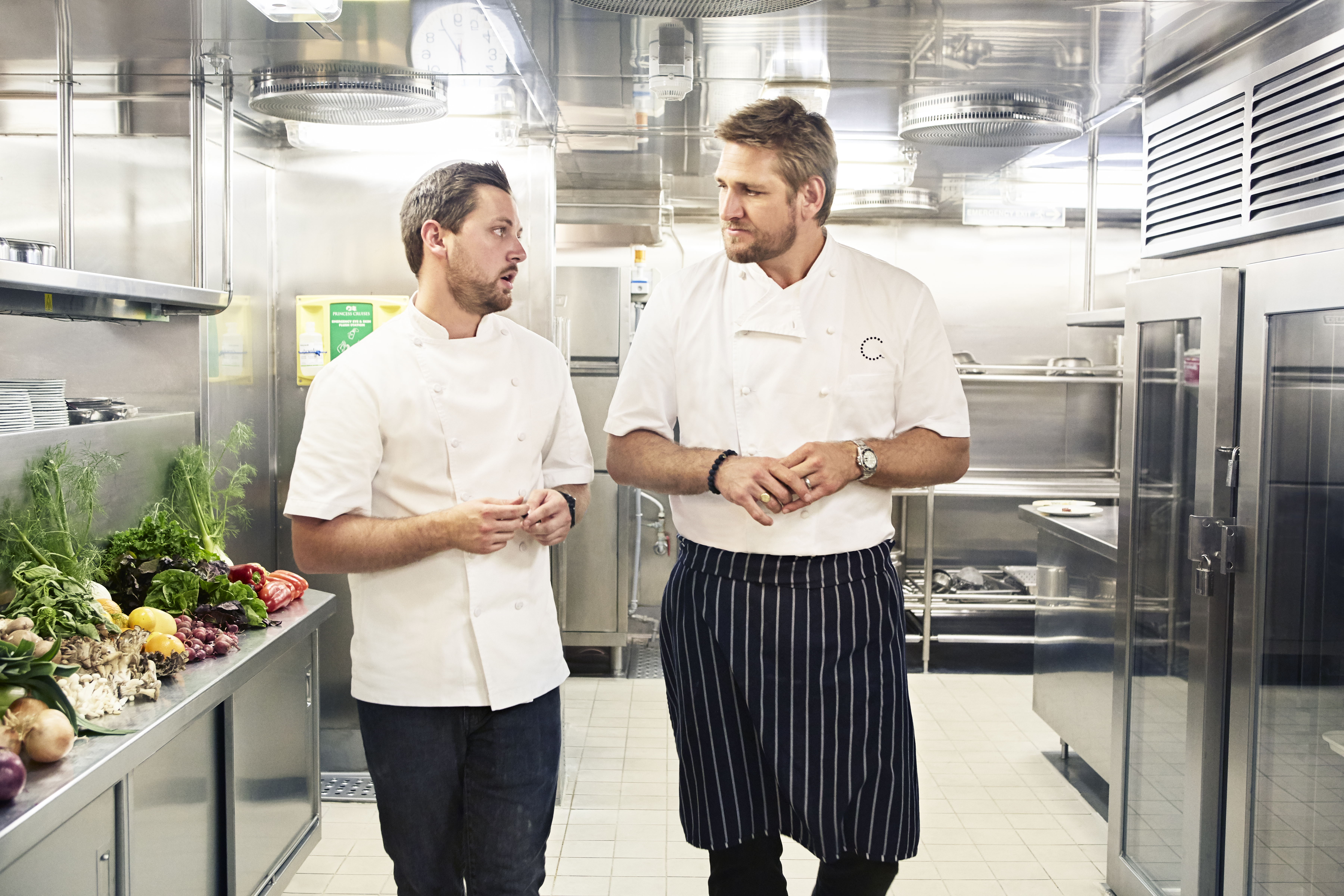 Curtis Stone and Christian Dortch talk while walking through a kitchen in chef’s clothes.