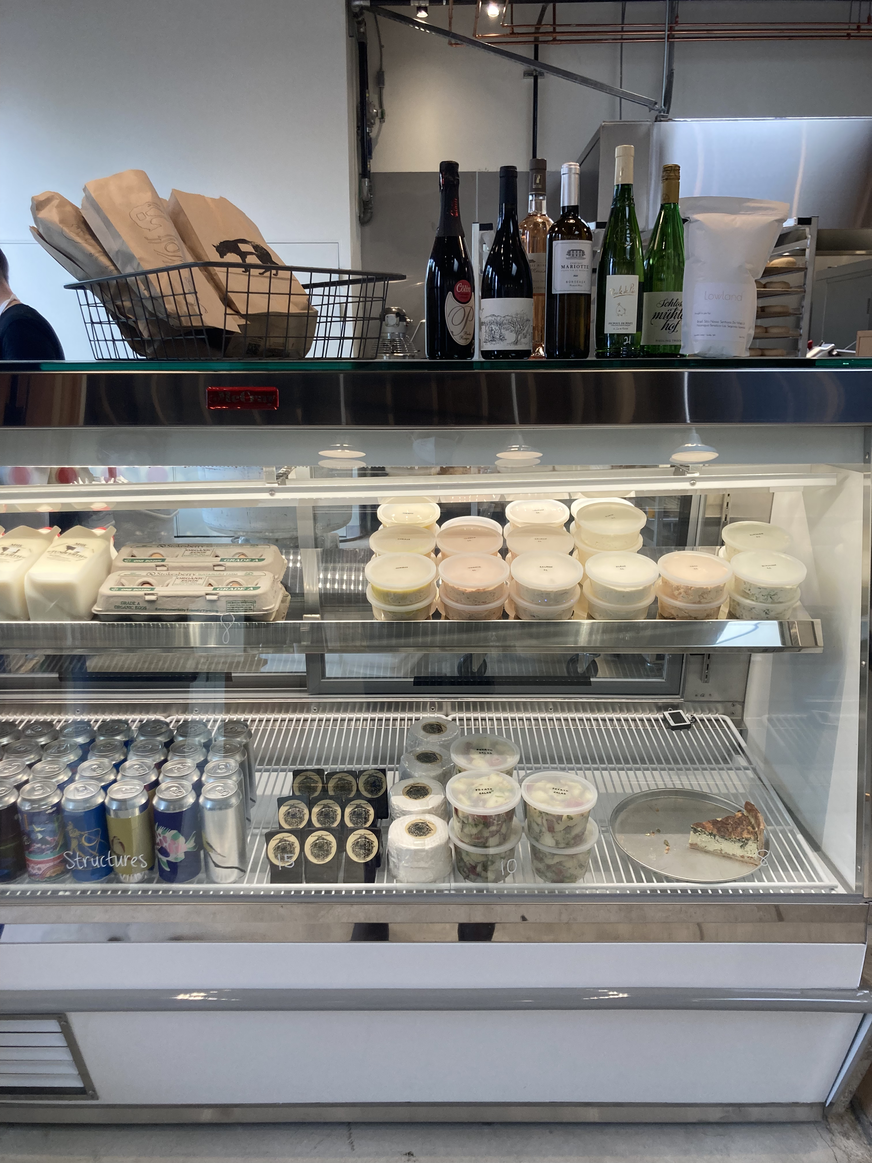 A deli case filled with cream cheese spreads in plastic containers, cans of beer, milk, cartons of eggs, cheese, and more.