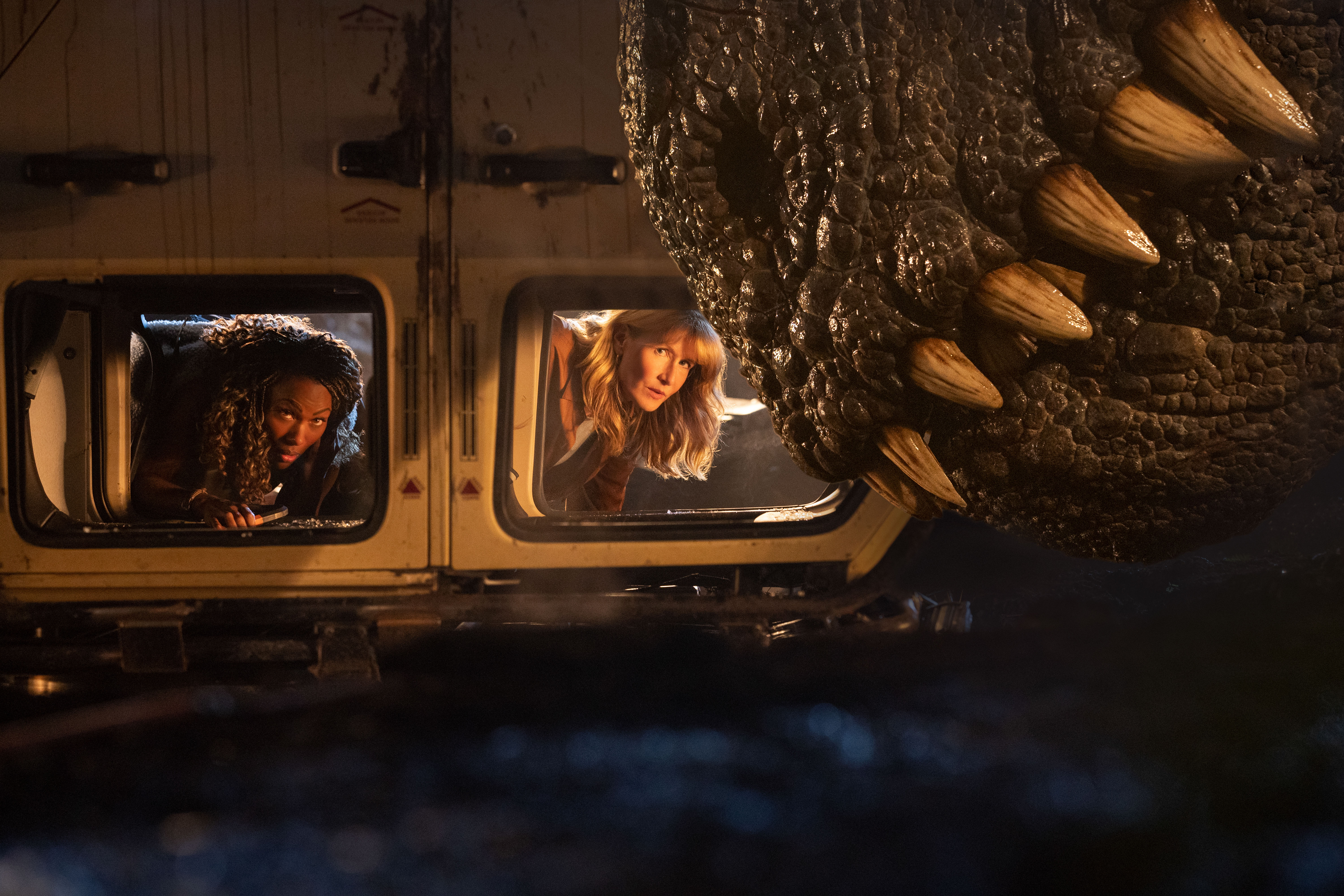 The snout of a large dinosaur is visible above the windows of a flipped-over car containing Laura Dern and DeWanda Wise.
