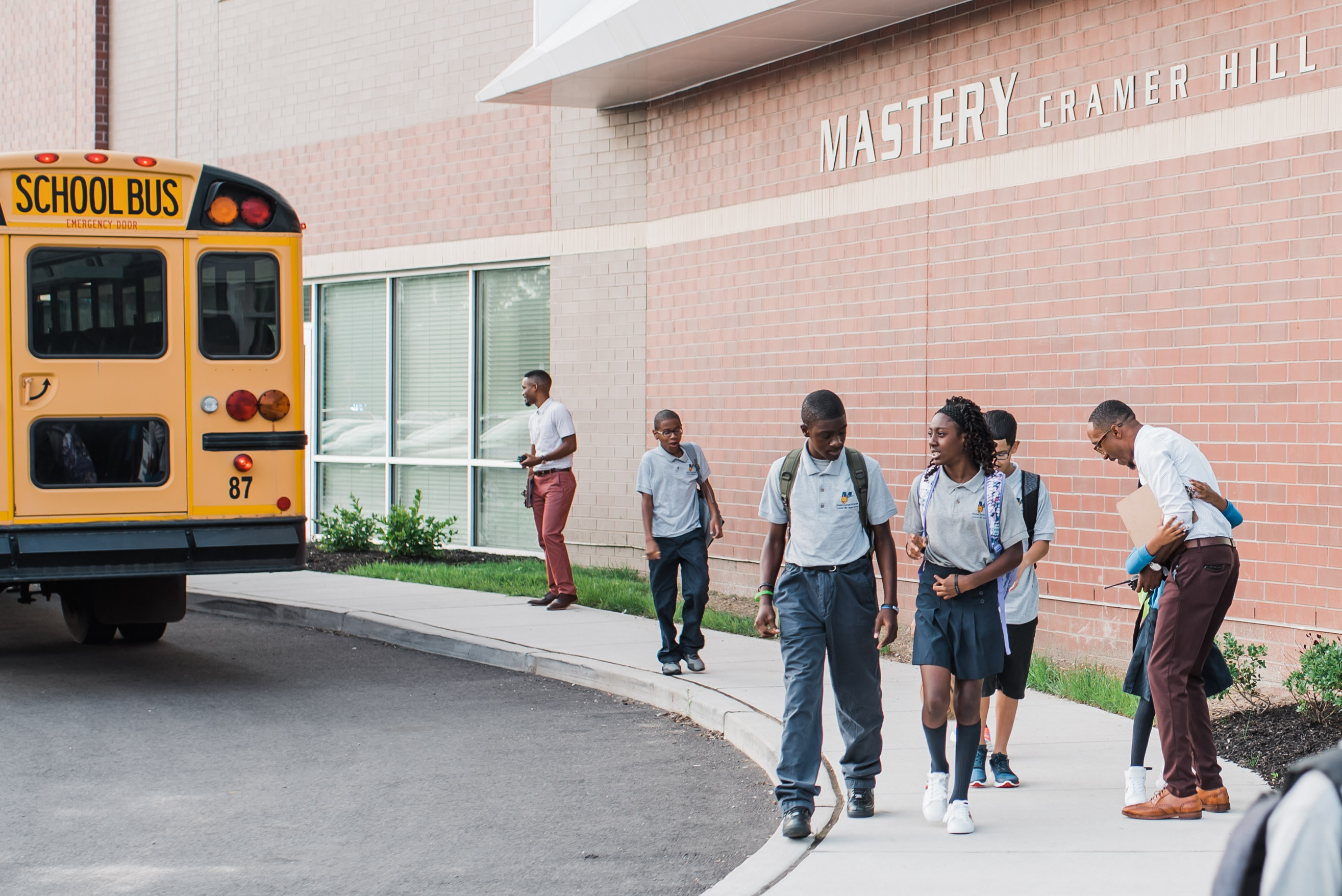 Students walk along the sidewalk beside a school bus in front of a school.