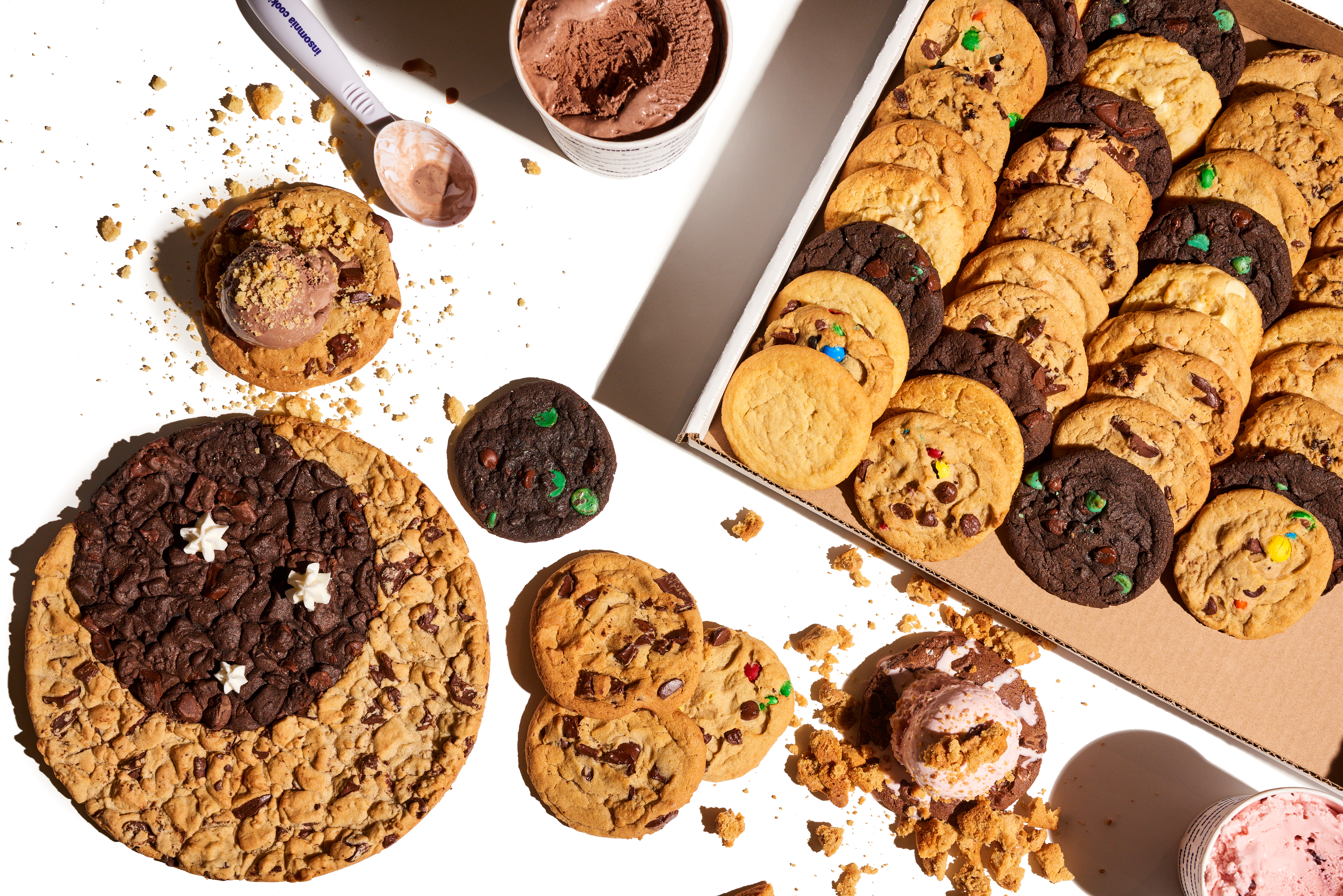 A cookie sheet and a table filled with an assortment of Insomnia cookies, plus a tub of chocolate ice cream.