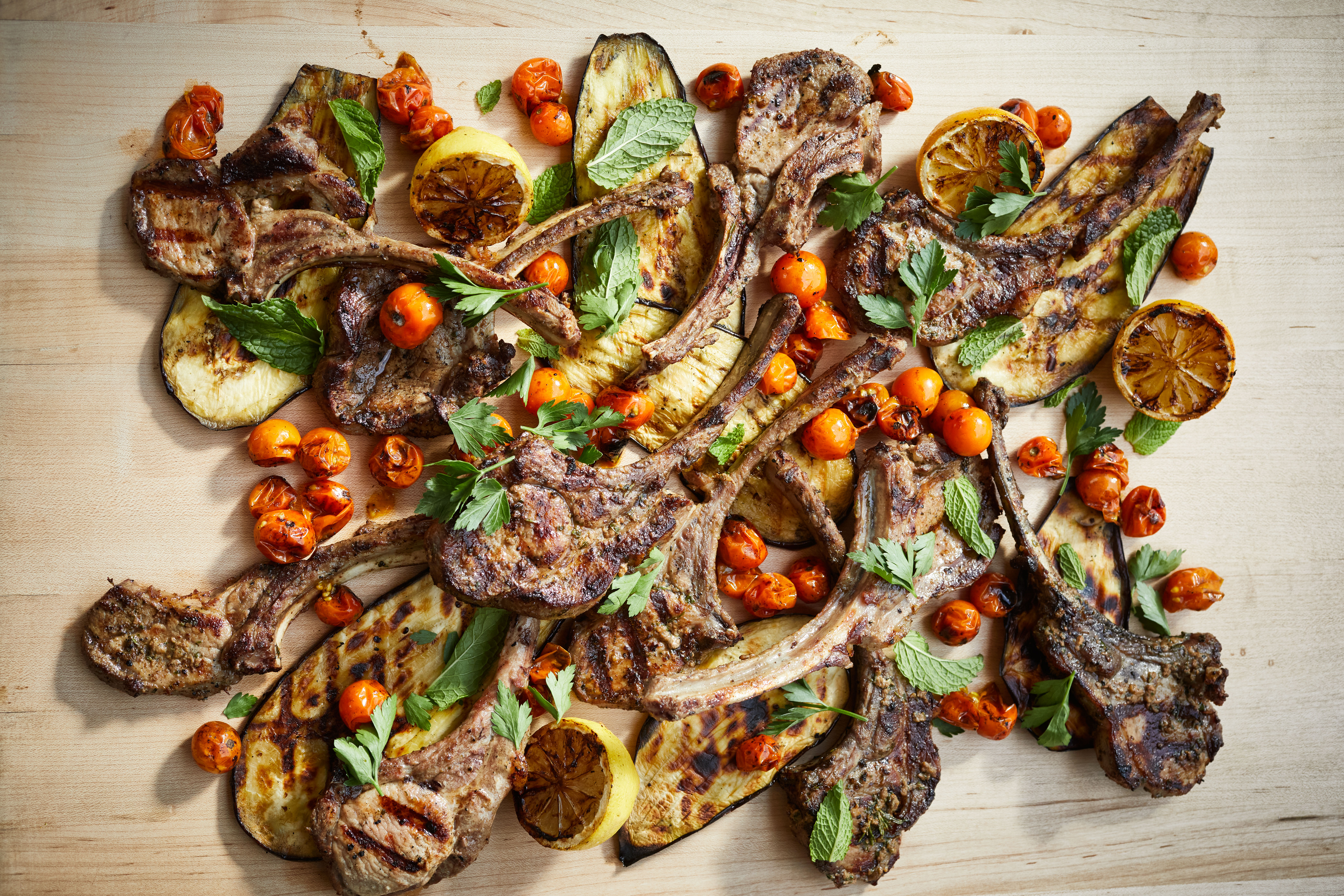 Several bone-in hog chops spread out on a light beige surface with long slices of roasted eggplant, roasted tomatoes, halved roasted lemons, leafy green herbs
