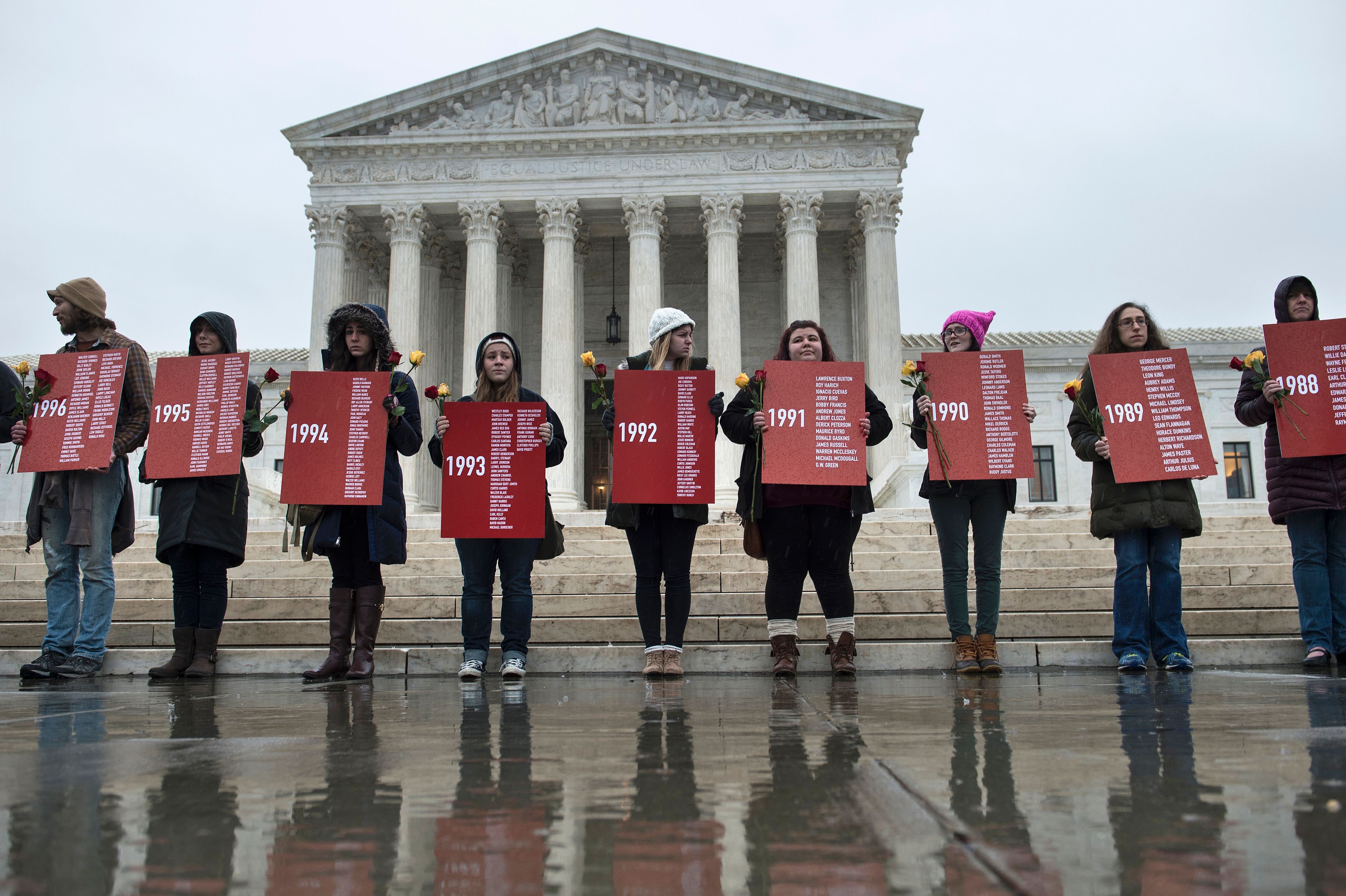 A line of people holding red signs stand in front of the US Supreme Court’s white marble steps. 