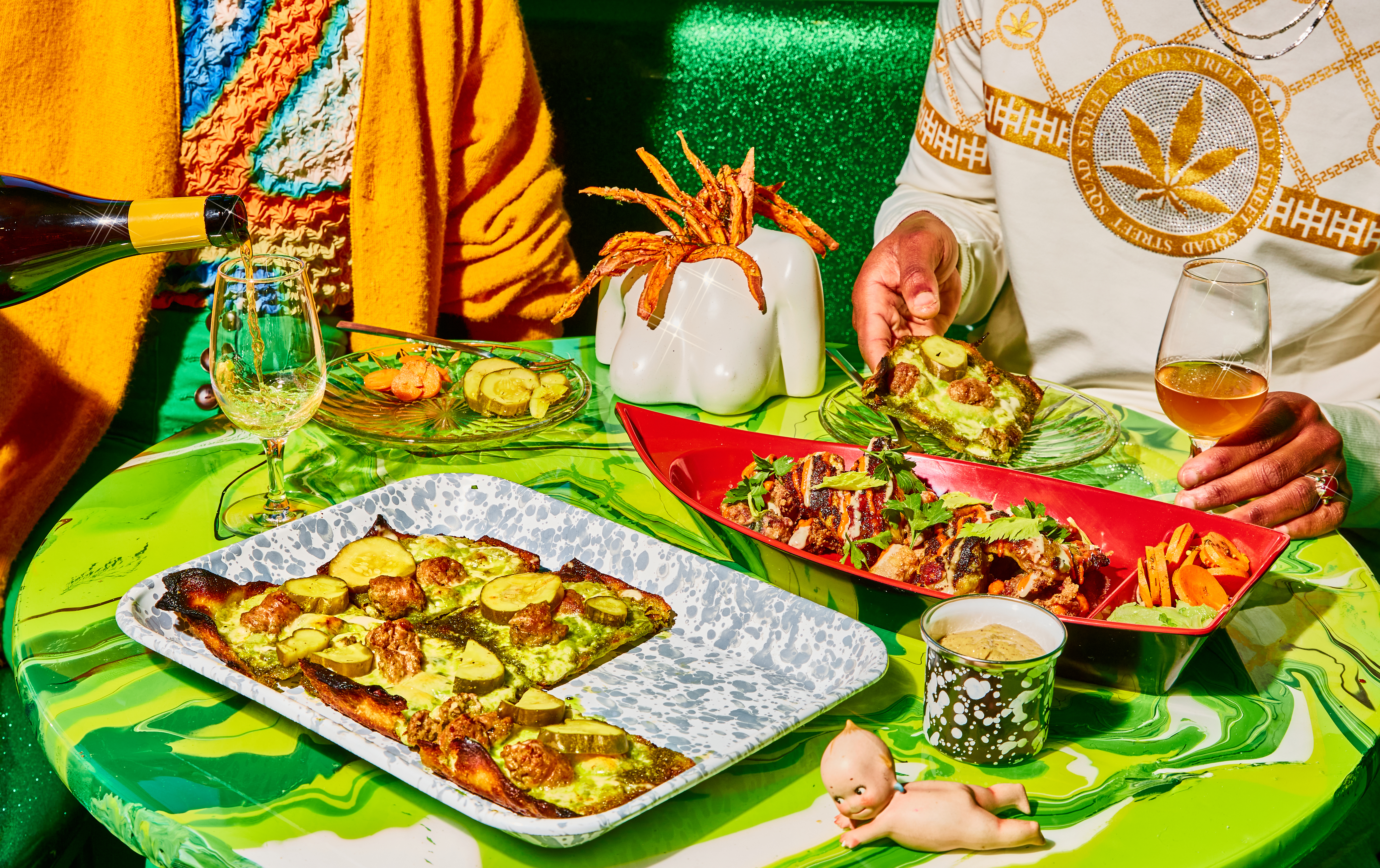 Diners sit in a bright green booth at a bright green swirled tabletop. On the table there’s a full rectangular pizza, a large red plastic boat full of grilled chicken, sweet potato fries served in a headless bust, and wine.