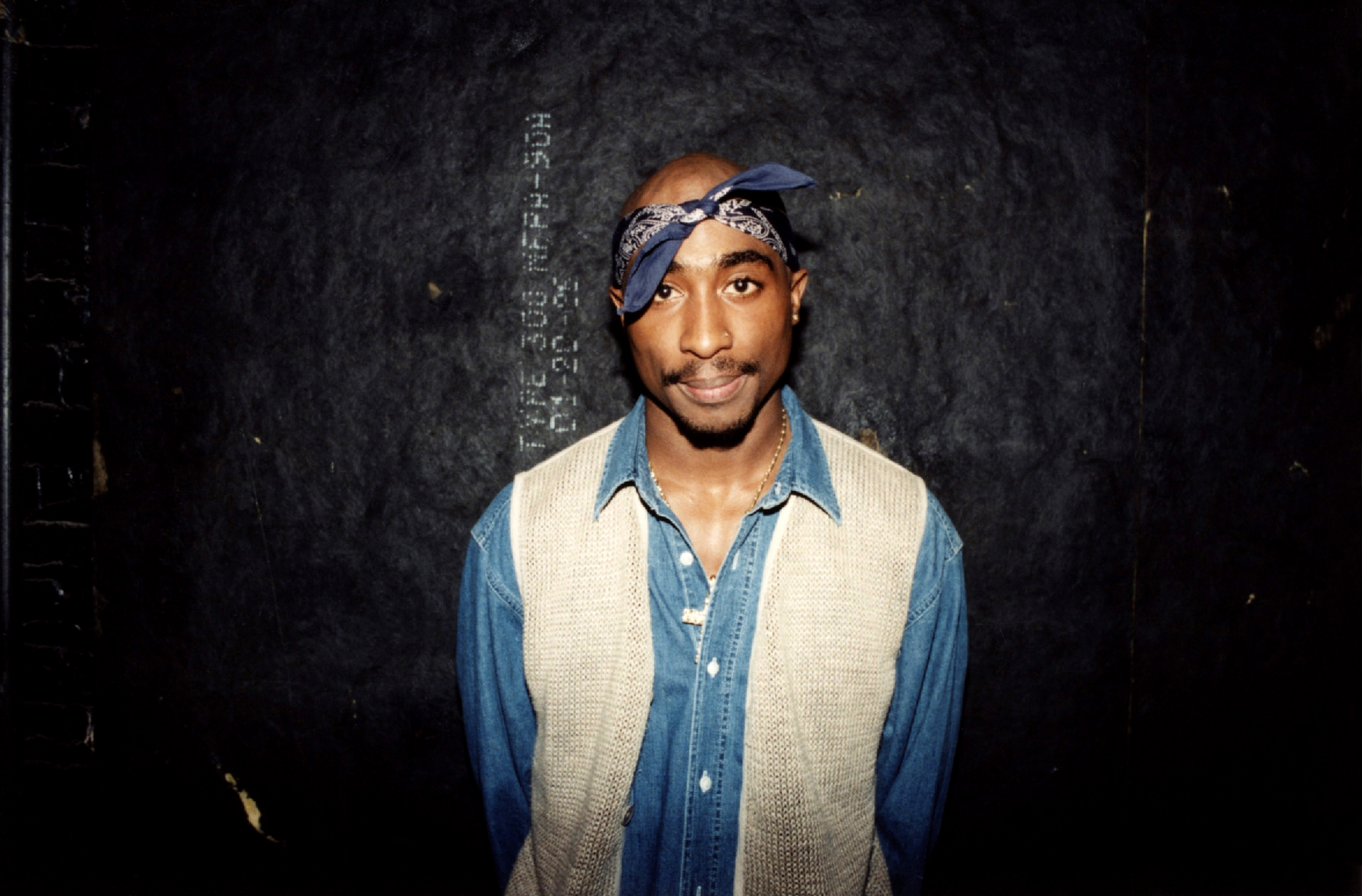 Tupac Shakur stands against a wall