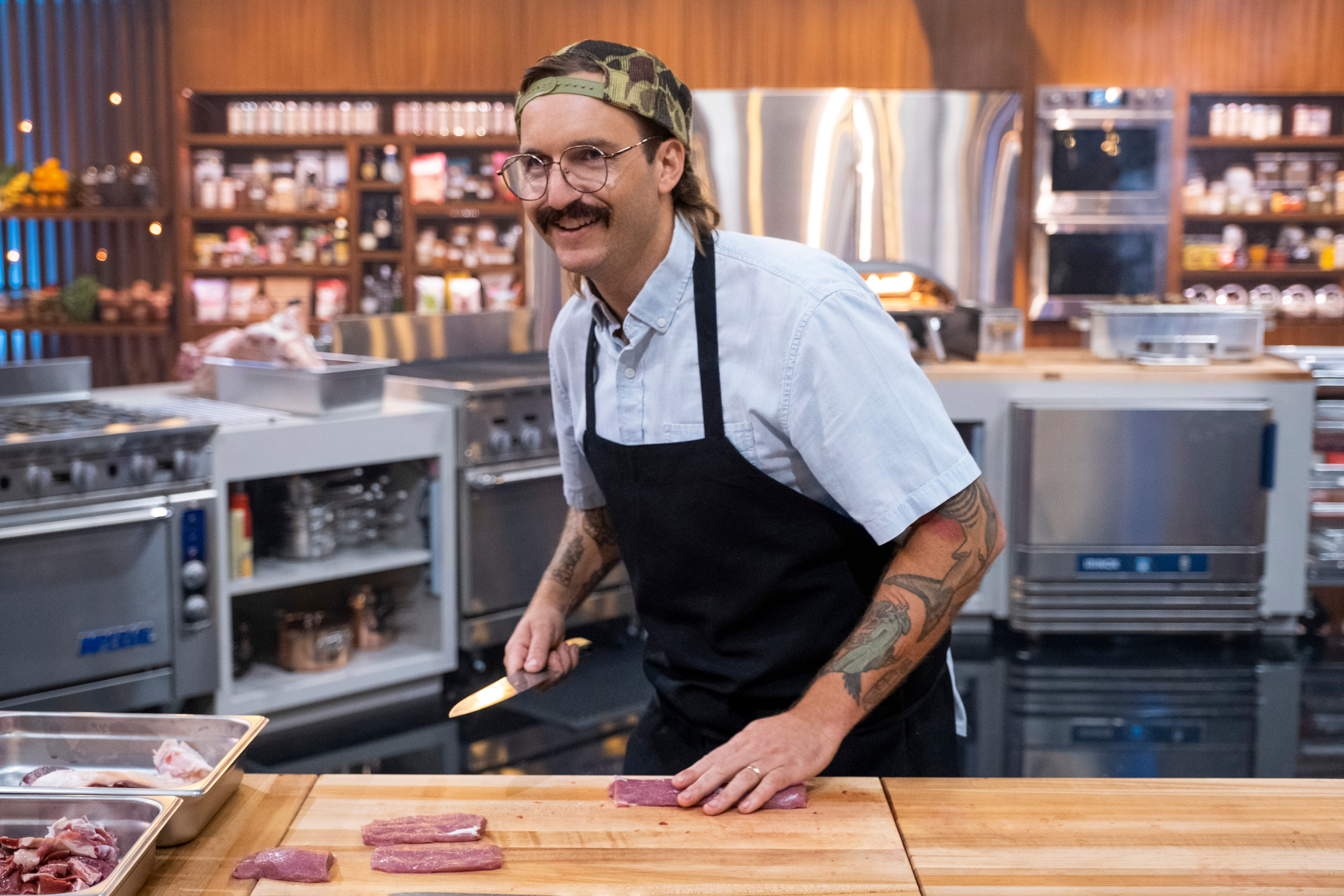 A white man wearing a backwards hat, glasses, and apron slices lamb meat thin at a counter in the Iron Chef Kitchen Stadium.