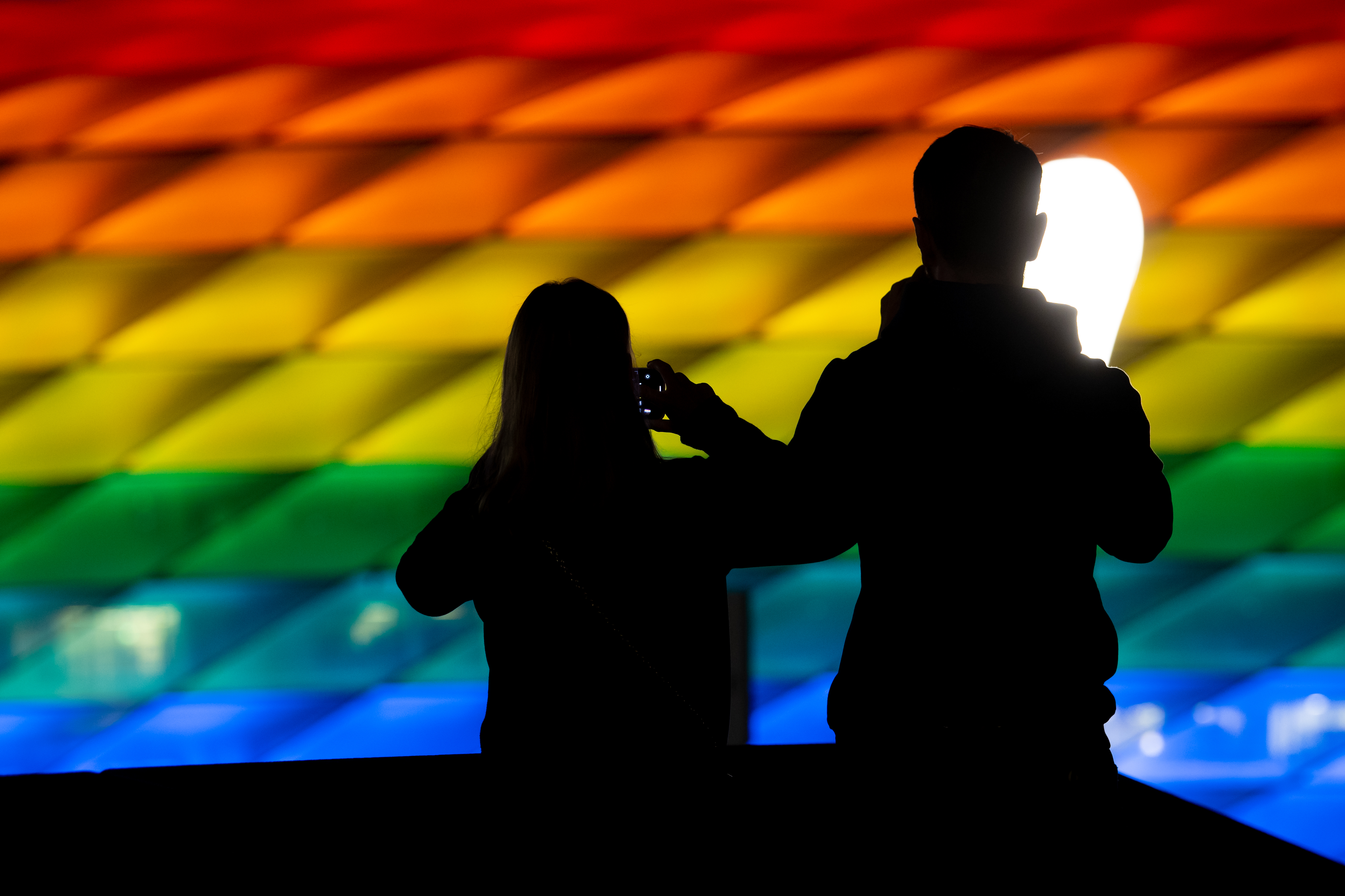 Two silhouettes in front of the rainbow-lit Allianz Arena at nighttime, taking photos