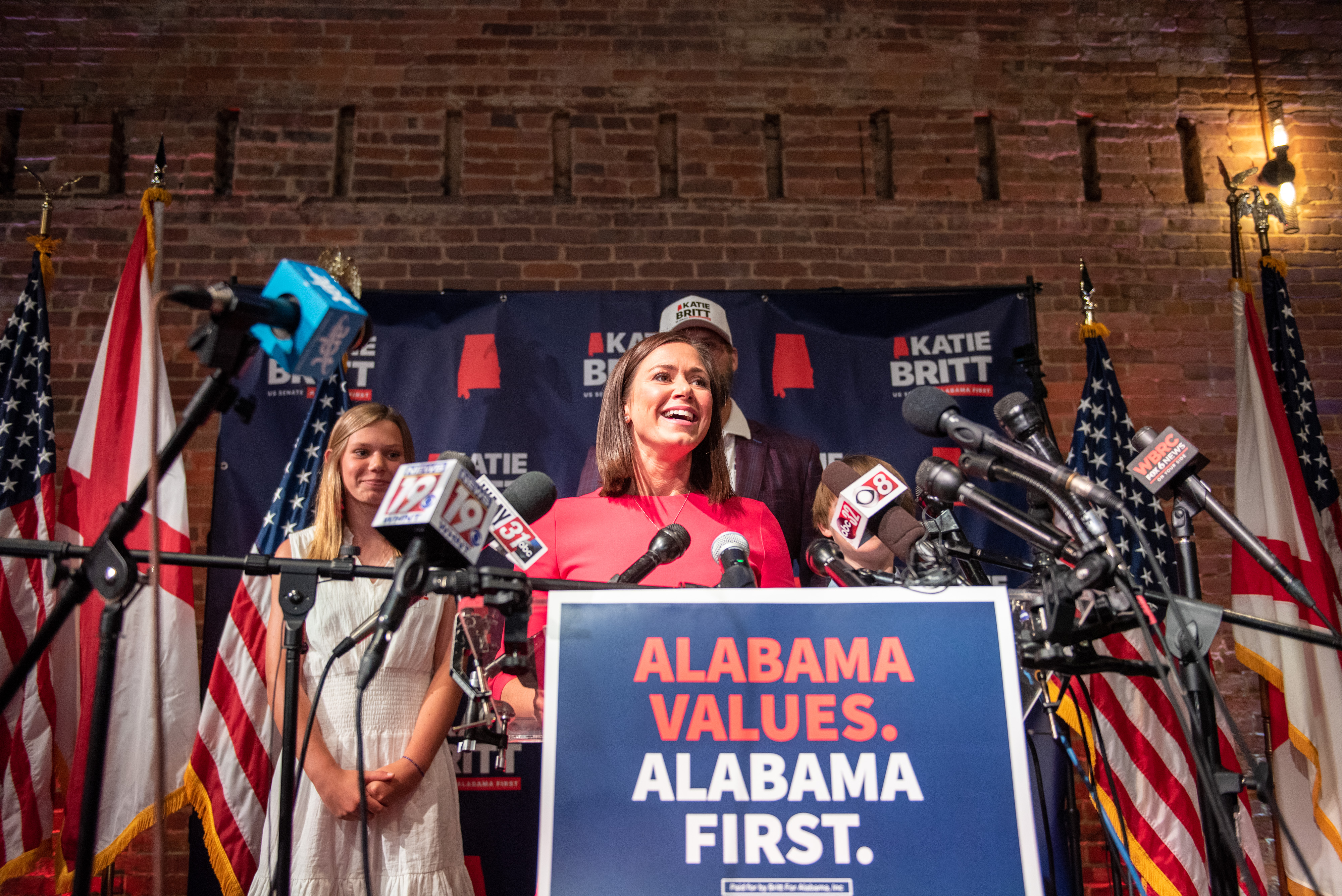 Britt, with a brown bob in a red dress, stands behind a podium emblazoned with the message: “Alabama Values. Alabama First.” Behind her are US and Alabama flags, as well as her family.