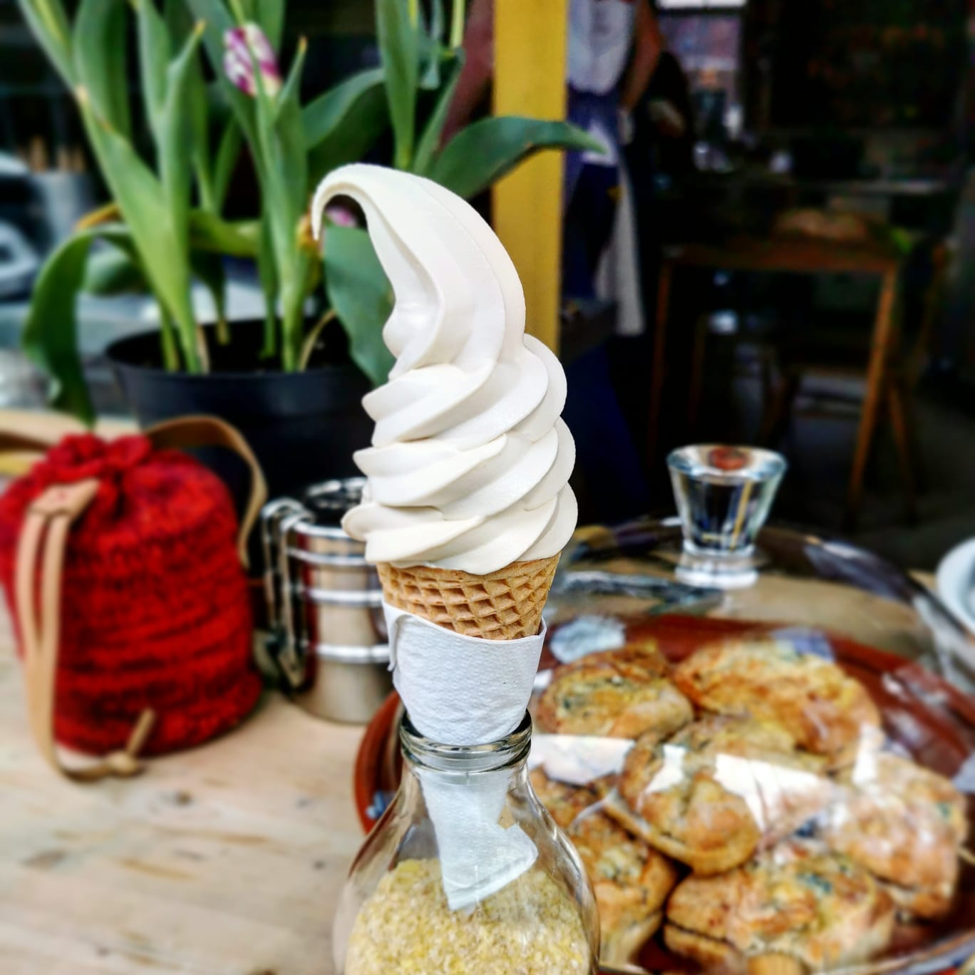 A cone of hazelnut soft serve sits in a glass bottle filled with sand, with pastries, a pot plant, and a red rattan fabric in the background