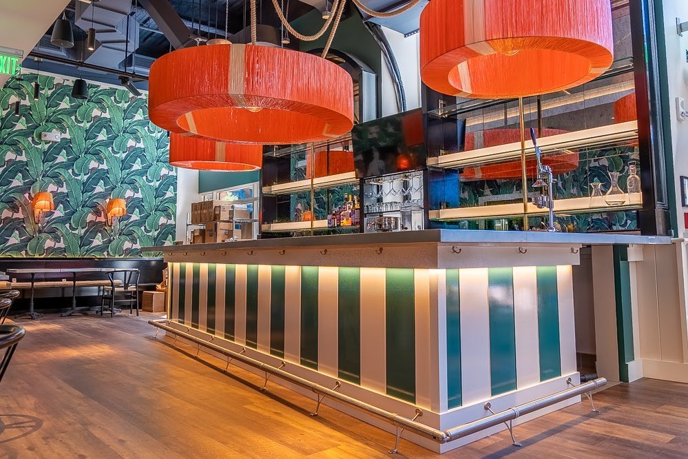 A restaurant interior features a bar with bold green and white stripes, green tropical leaf wallpaper, and large orange light fixtures. 