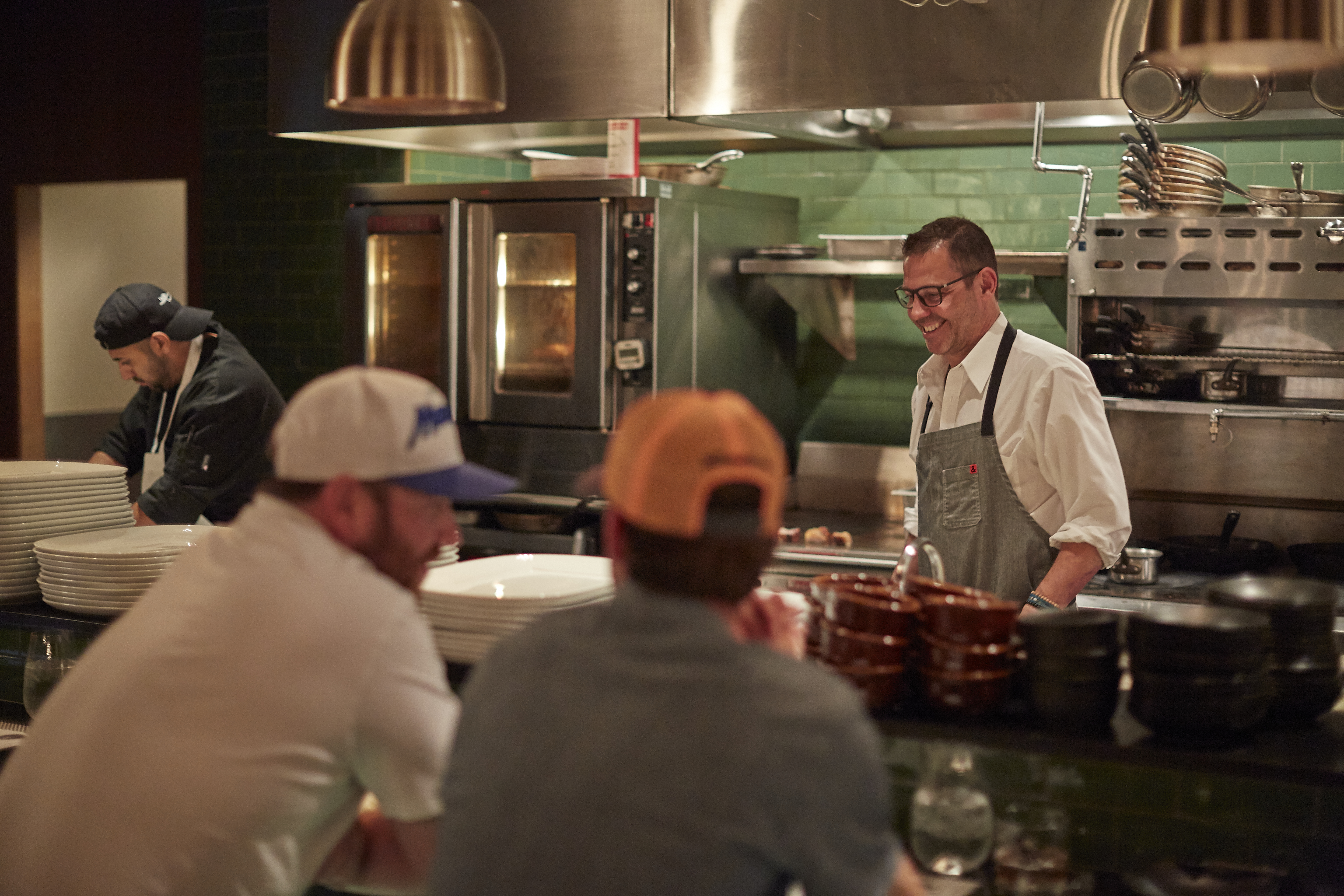 Diners in baseball caps sit in the foreground while chef John Tesar laughs in the kitchen of Knife in the background. Behind him are the restaurant’s green tiled walls and steel kitchen appliances.