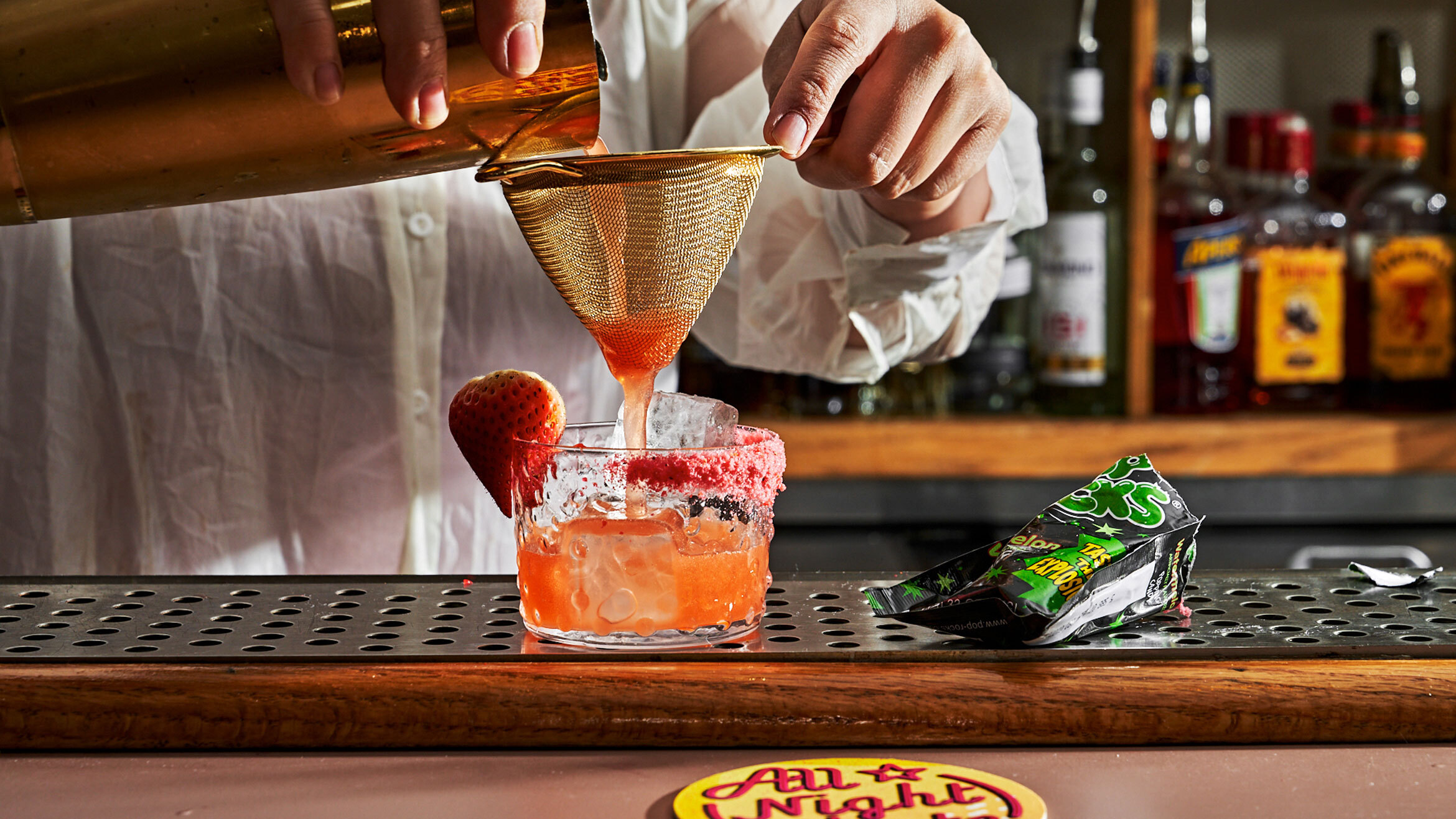 A bartender pours a drink through a strainer and into a cocktail glass with a Pop Rocks crusted rim.