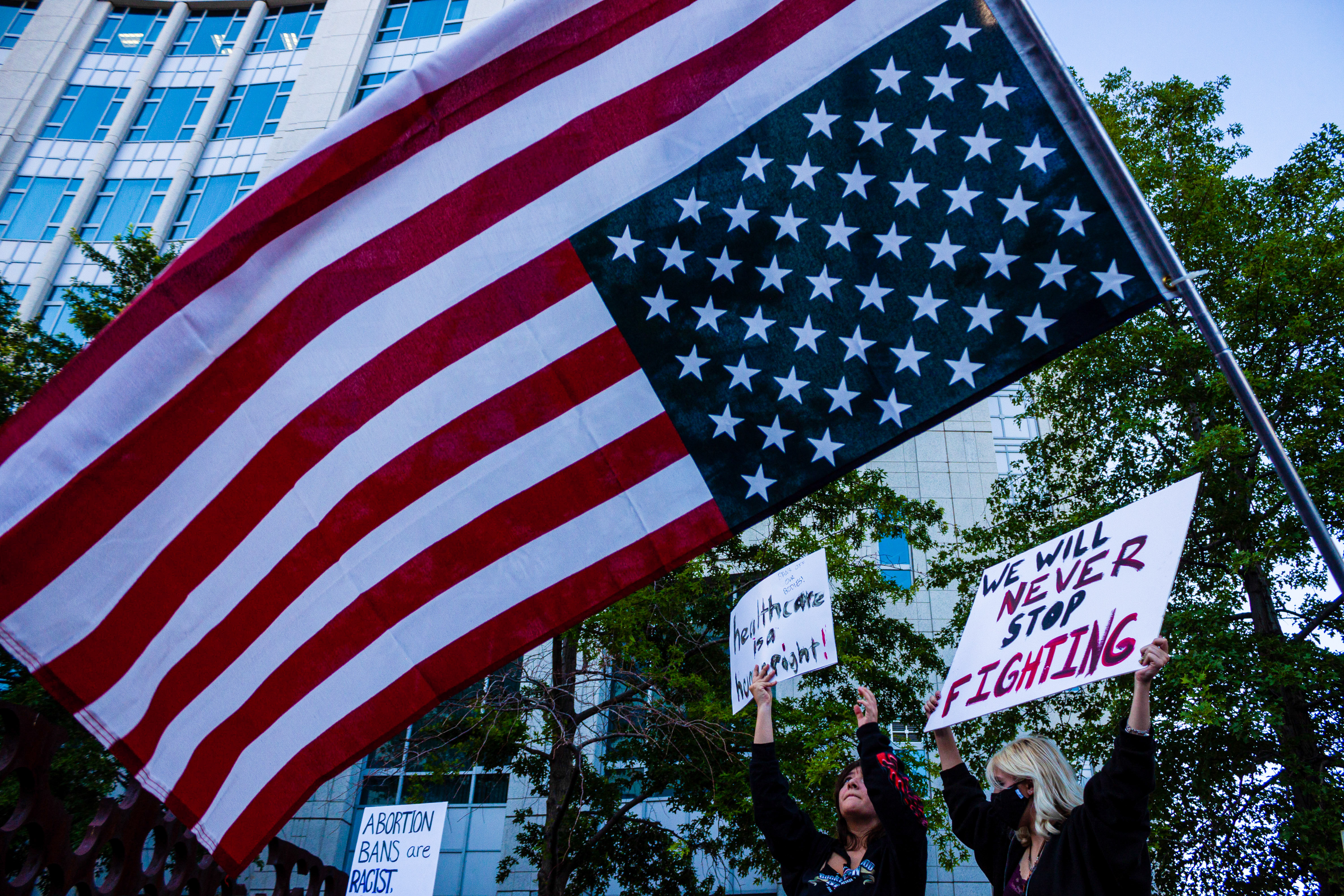 An upside-down American flag and signs including one reading “We will never stop fighting” amid a crowd of protesters in a city street.
