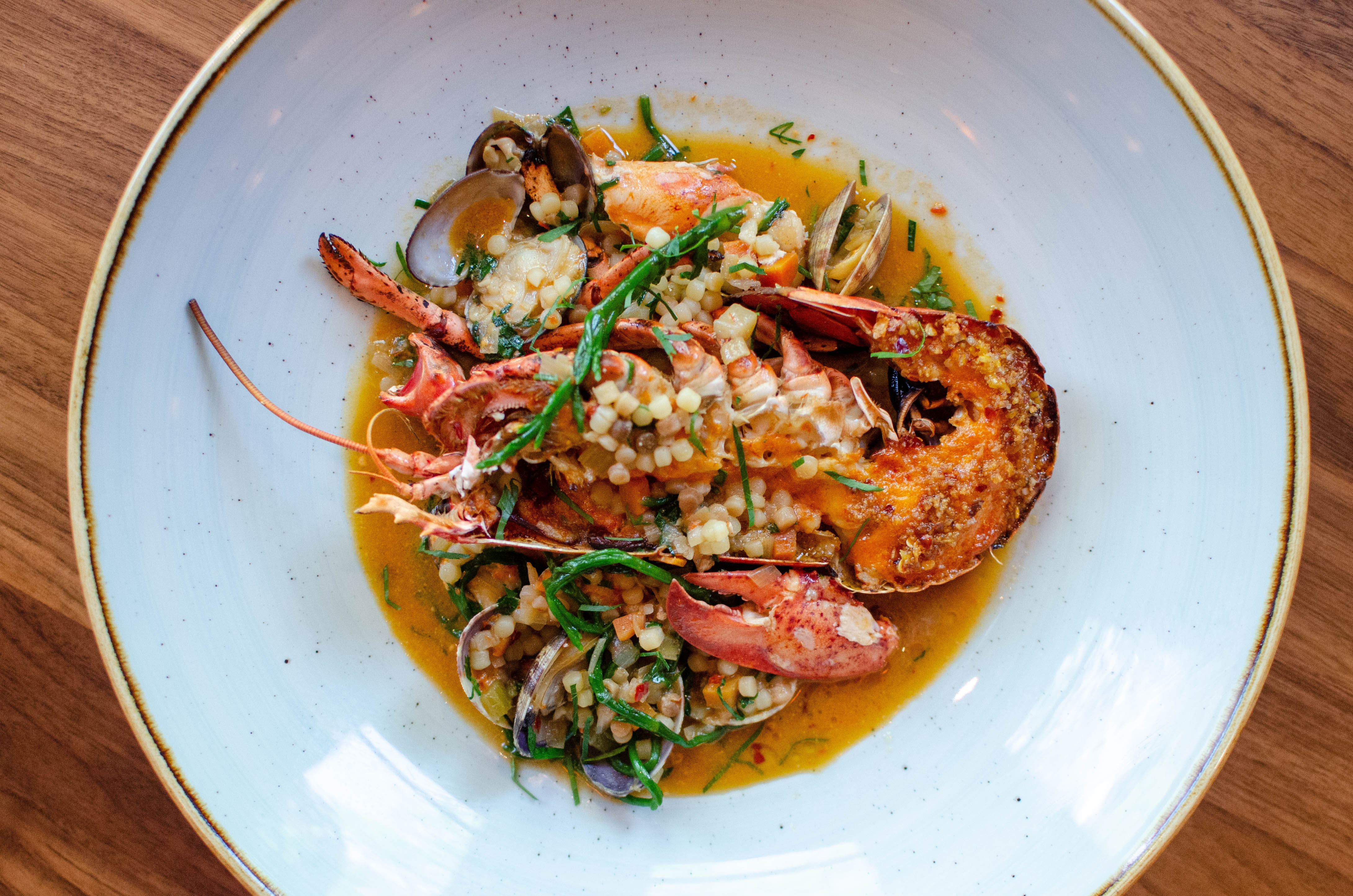 Overhead view of a half lobster sitting in a small pool of reddish broth and topped with small, round dots of pasta, herbs, and small clams.