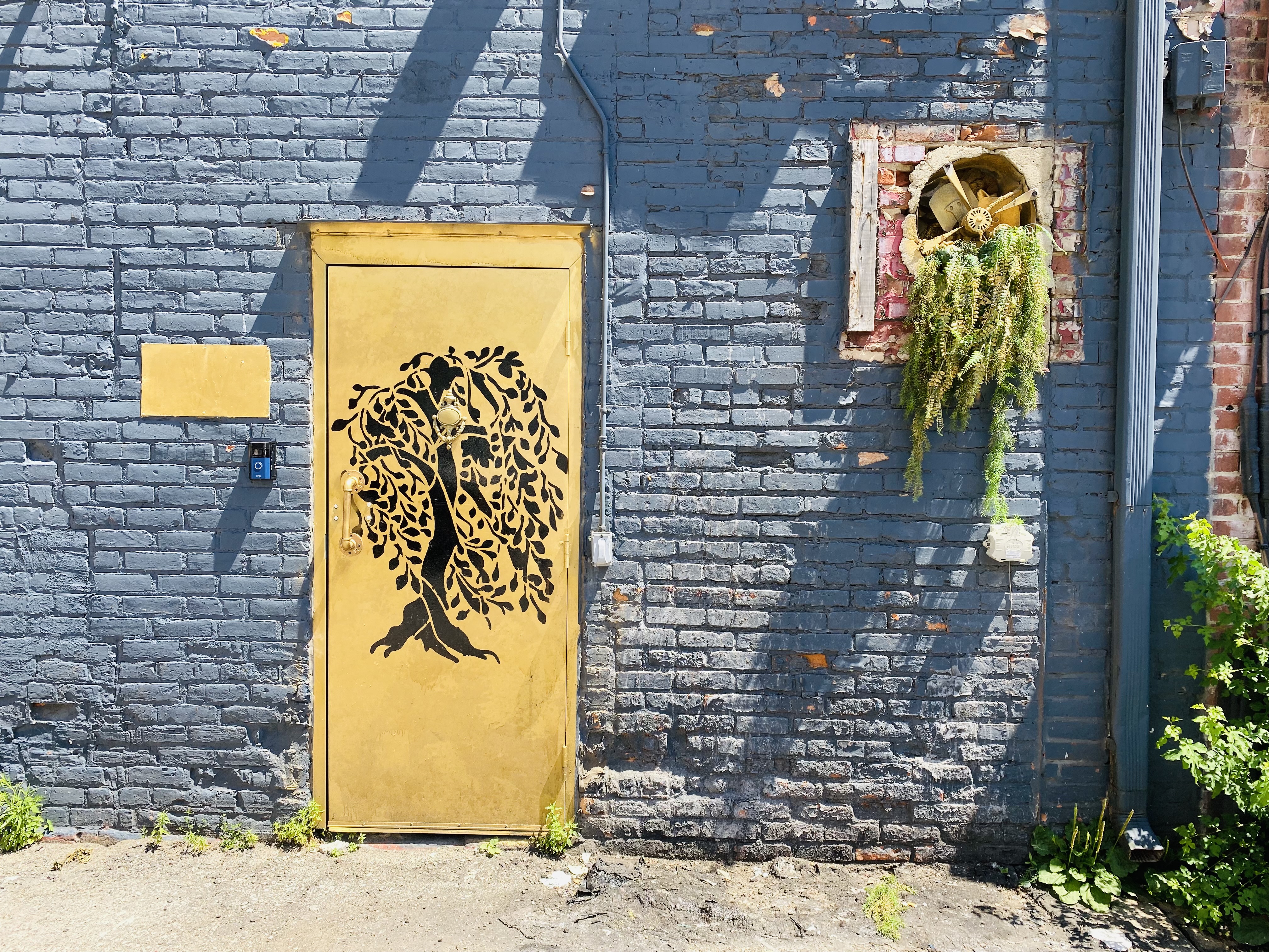 A golden door with a black willow tree silhouette painted on it against a blue gray brick wall in an alley in downtown Detroit
