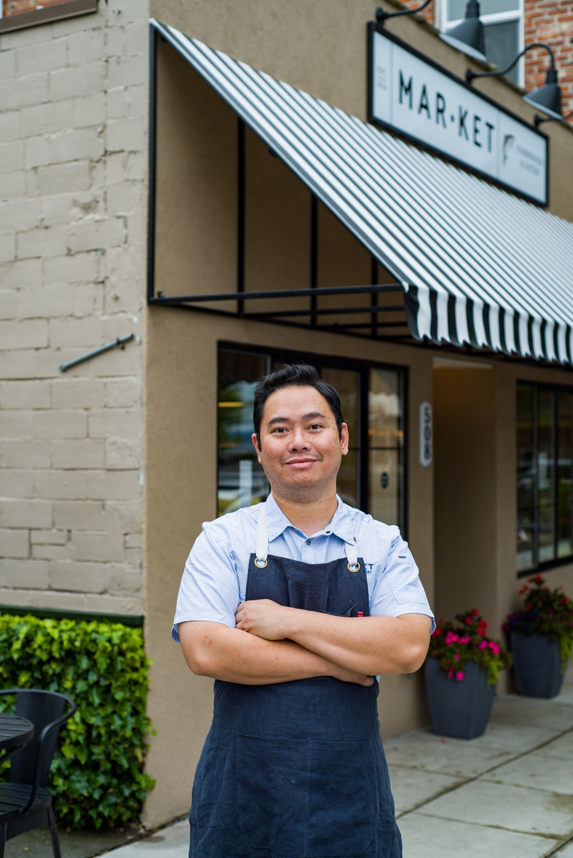 A man with medium-length black hair, wearing a black apron and a light blue button-down shirt, stands outside of a restaurant.