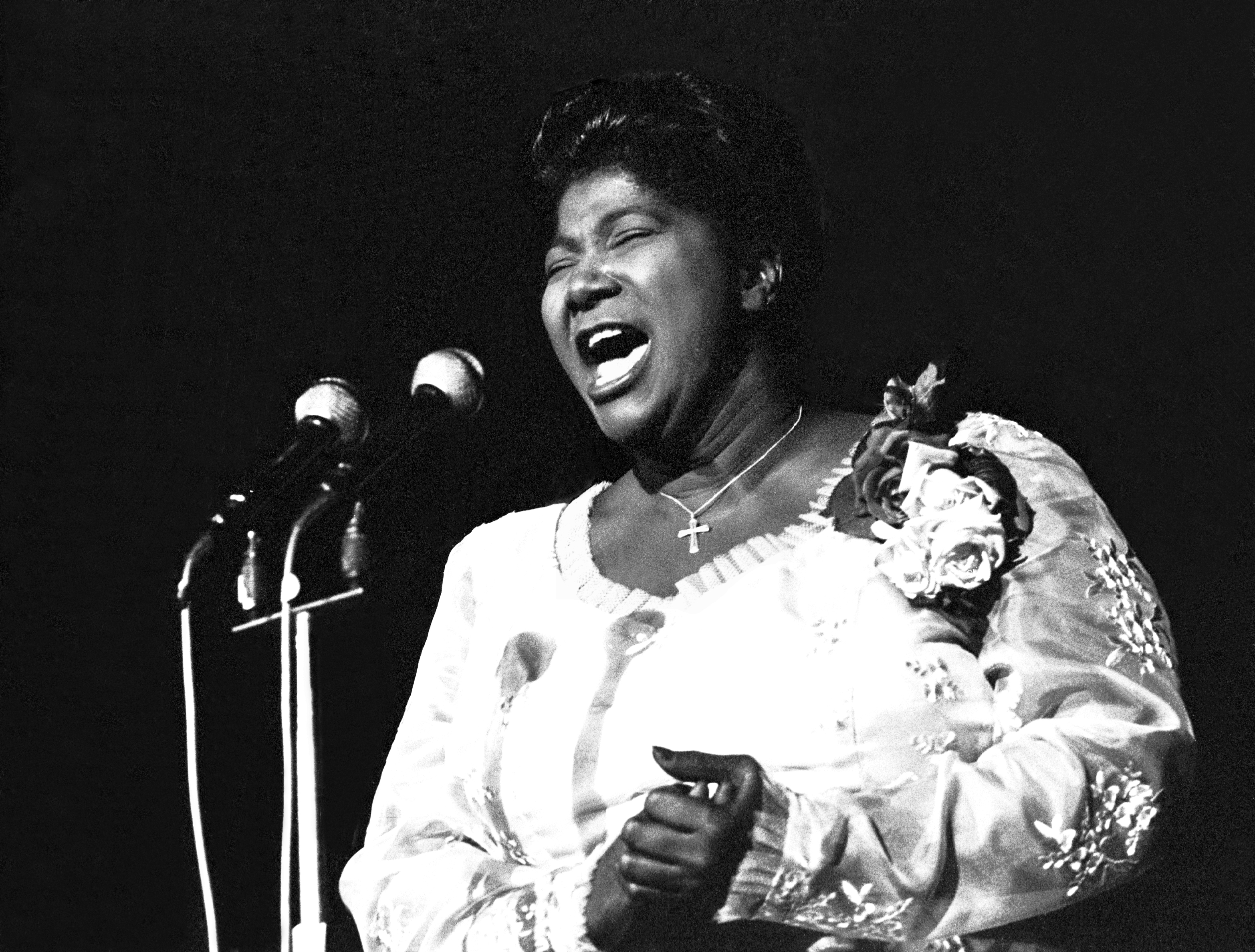 A black and white photo of gospel singer Mahalia Jackson, wearing an embroidered, light-colored, long-sleeve evening gown with a silk floral corsage, and, eyes closed, singing into two microphones against a black curtain.