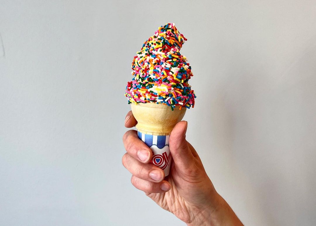 A hand holds up a cone of vanilla soft serve covered with chocolate sprinkles against a plain white background.