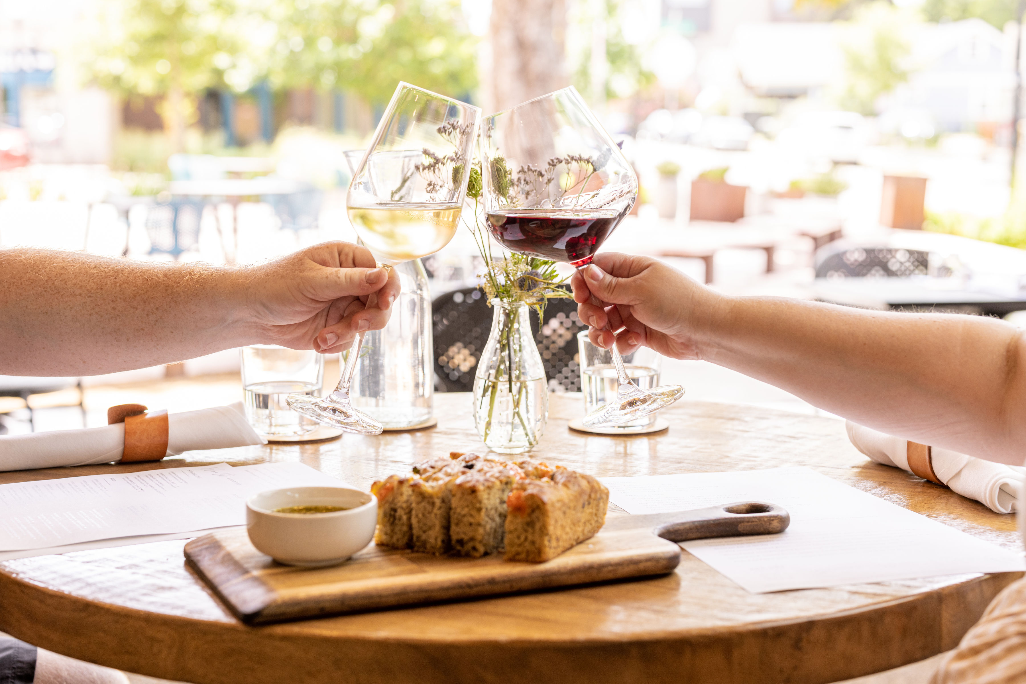 A pair of hands cheers with two wine glasses over a table with bread and other accouterment. 