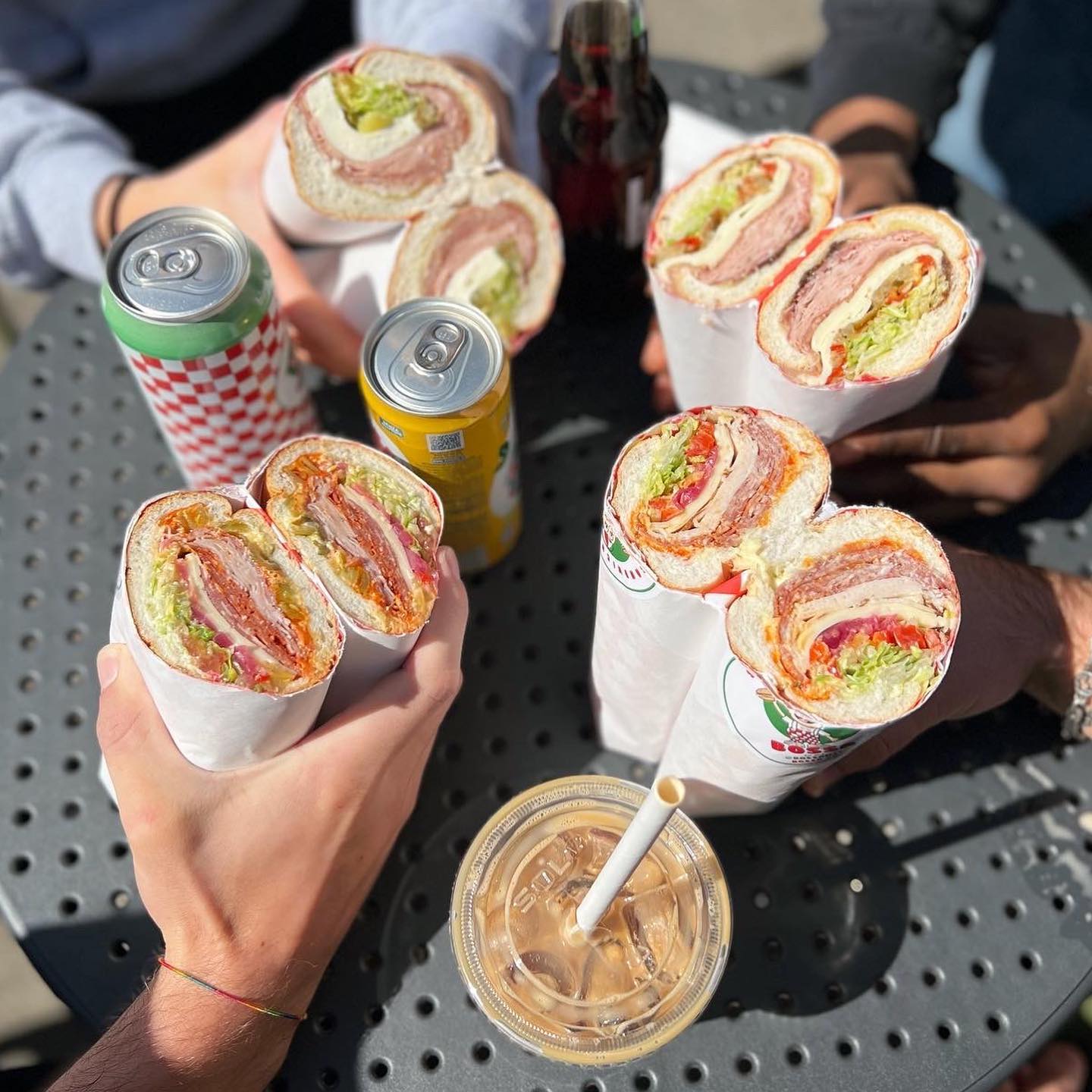 four people each holding a sandwich with two drink cans on table