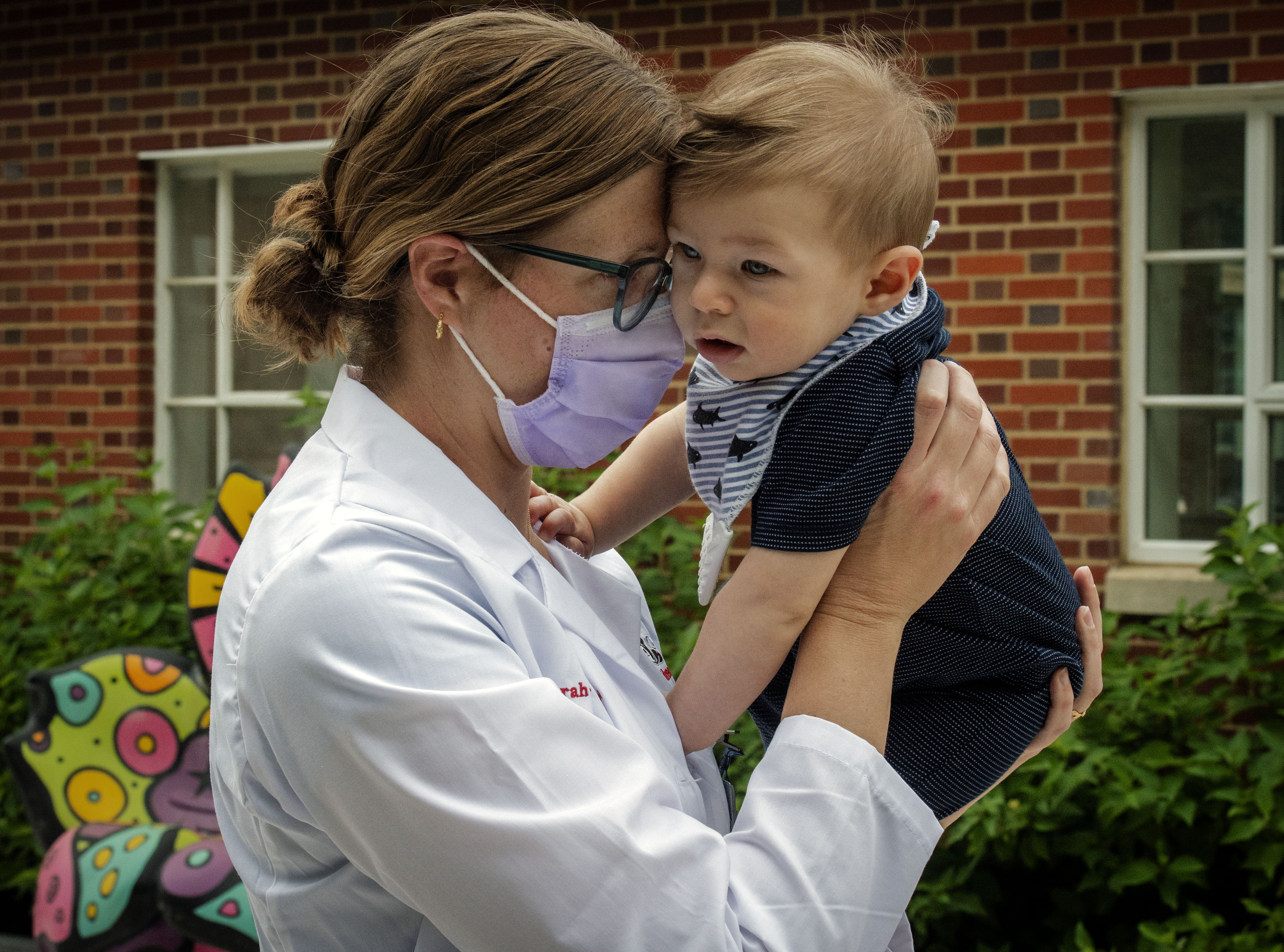 A doctor wearing a mask holding a baby outside a house.