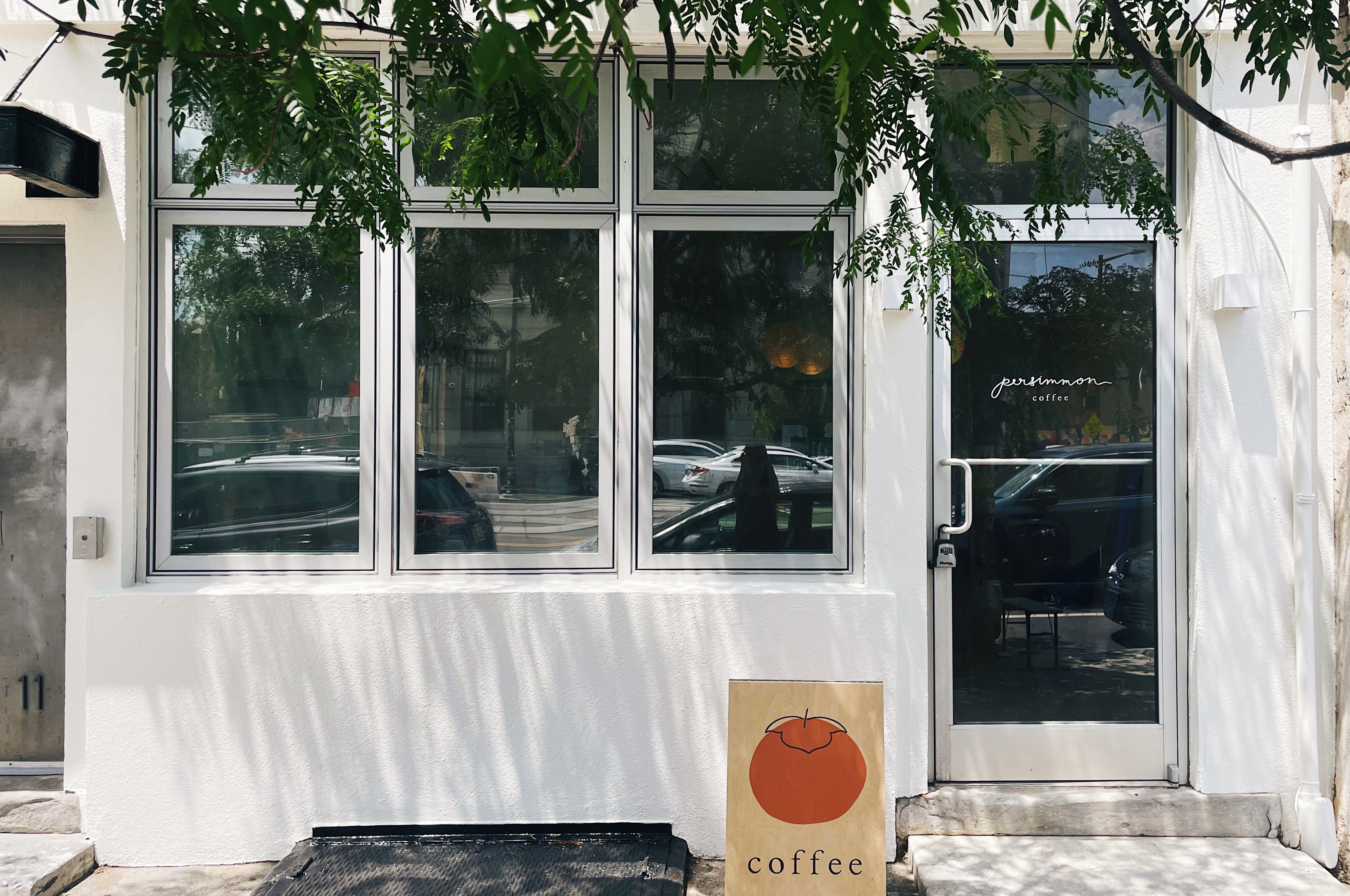 The outside of a shop with a white facade with a green tree and a sandwich board with an orange persimmon that reads Coffee.