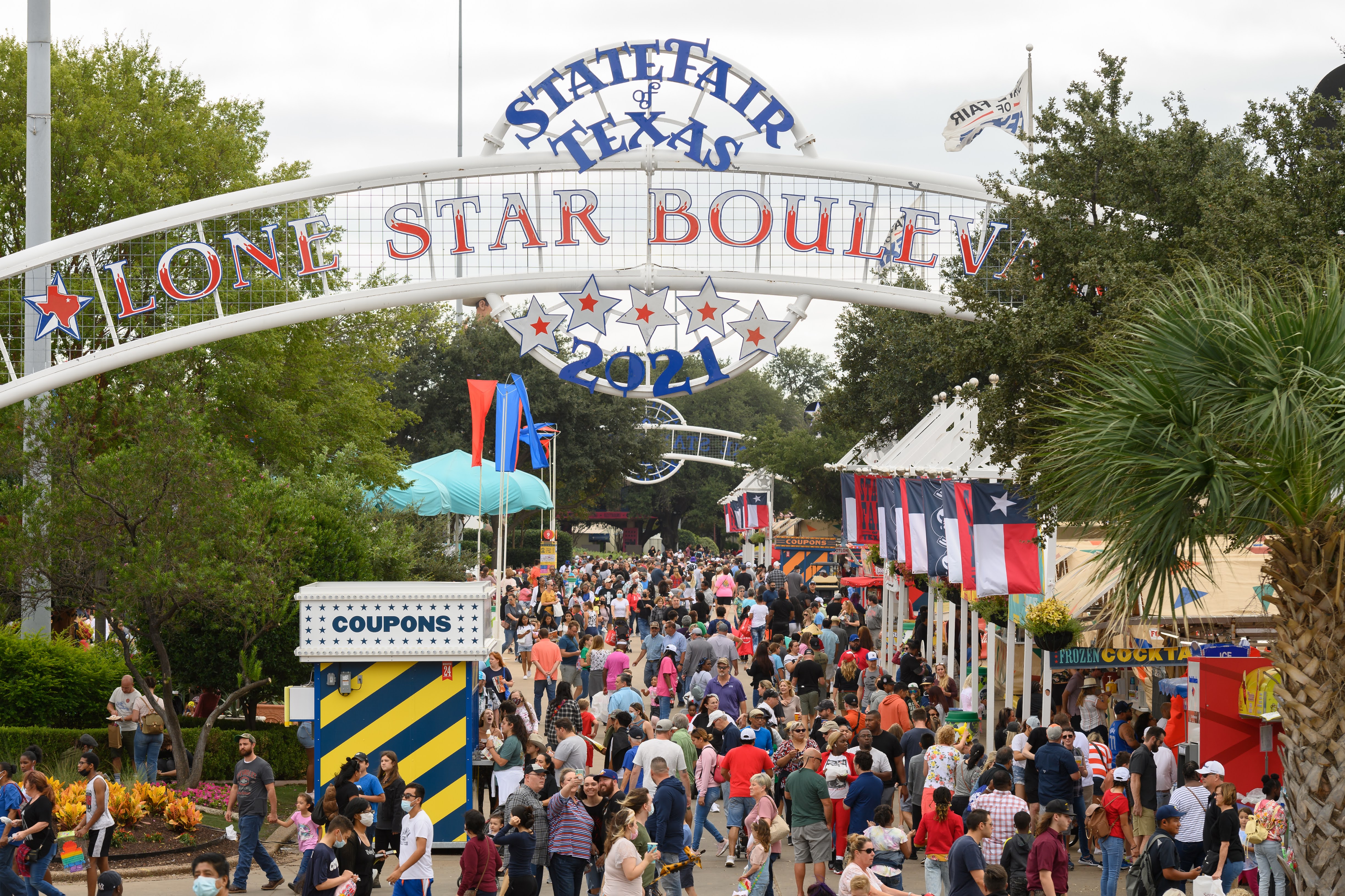 A parkway is full of people and each side is lined with food and ticket vendor booths. Above the scene, a metal overhang declares it, “State Fair of Texas, Lone Star Parkway.”