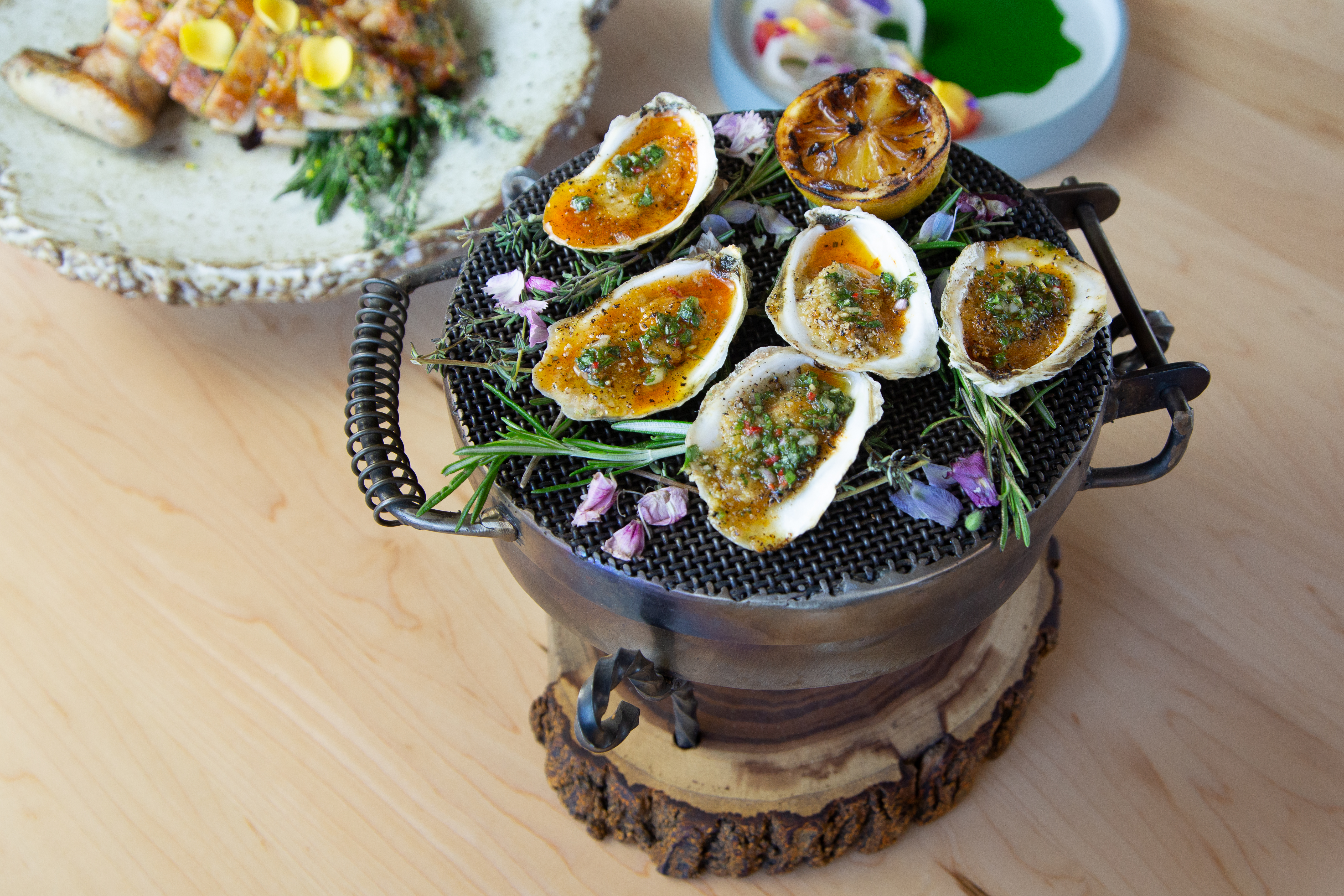 A cauldron of green-onion topped grilled oysters on the half shell, sitting on a light wood table among a couple other dishes of food.