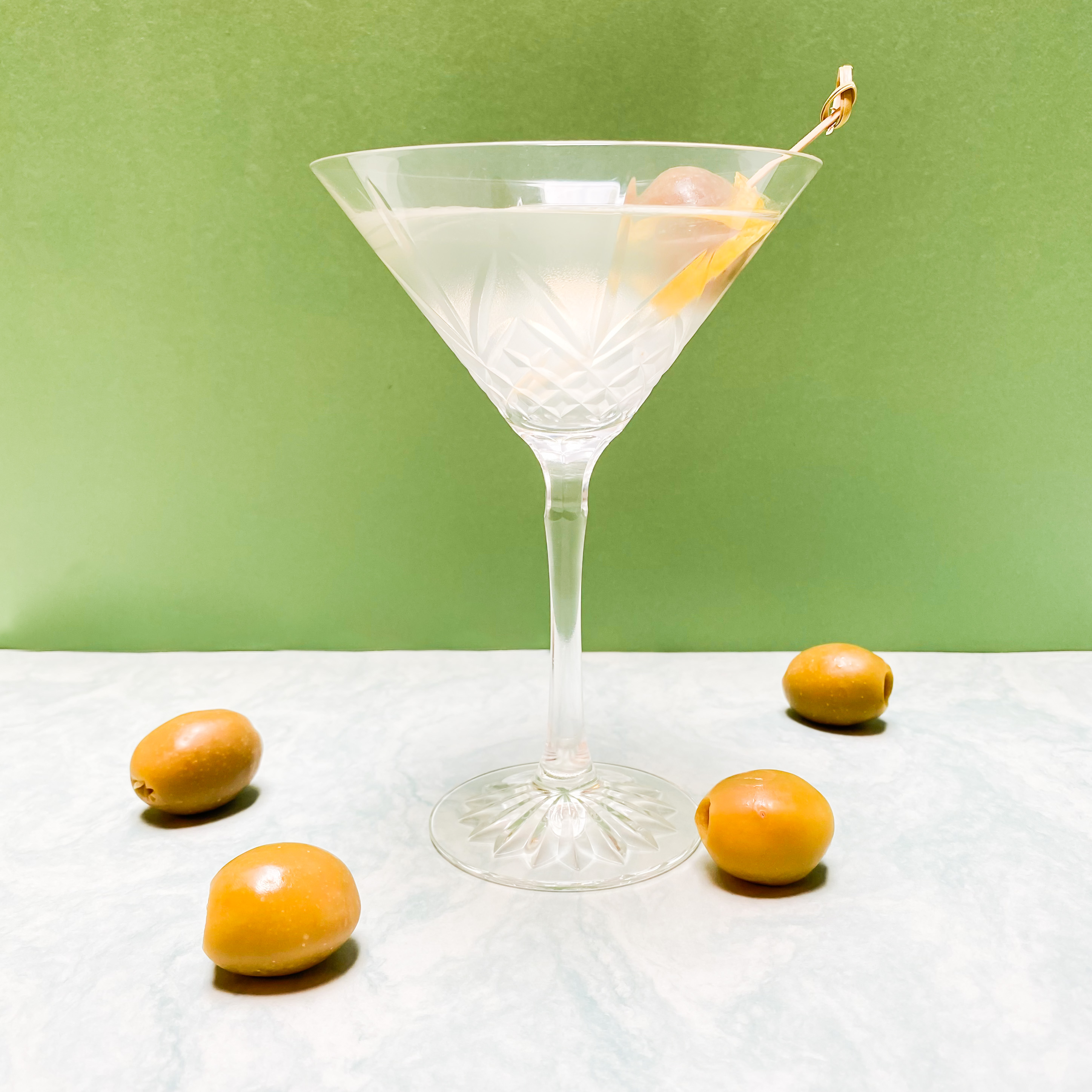 A clear drink in a martini glass pictured against a green background. A few olives are placed around the base of the glass.
